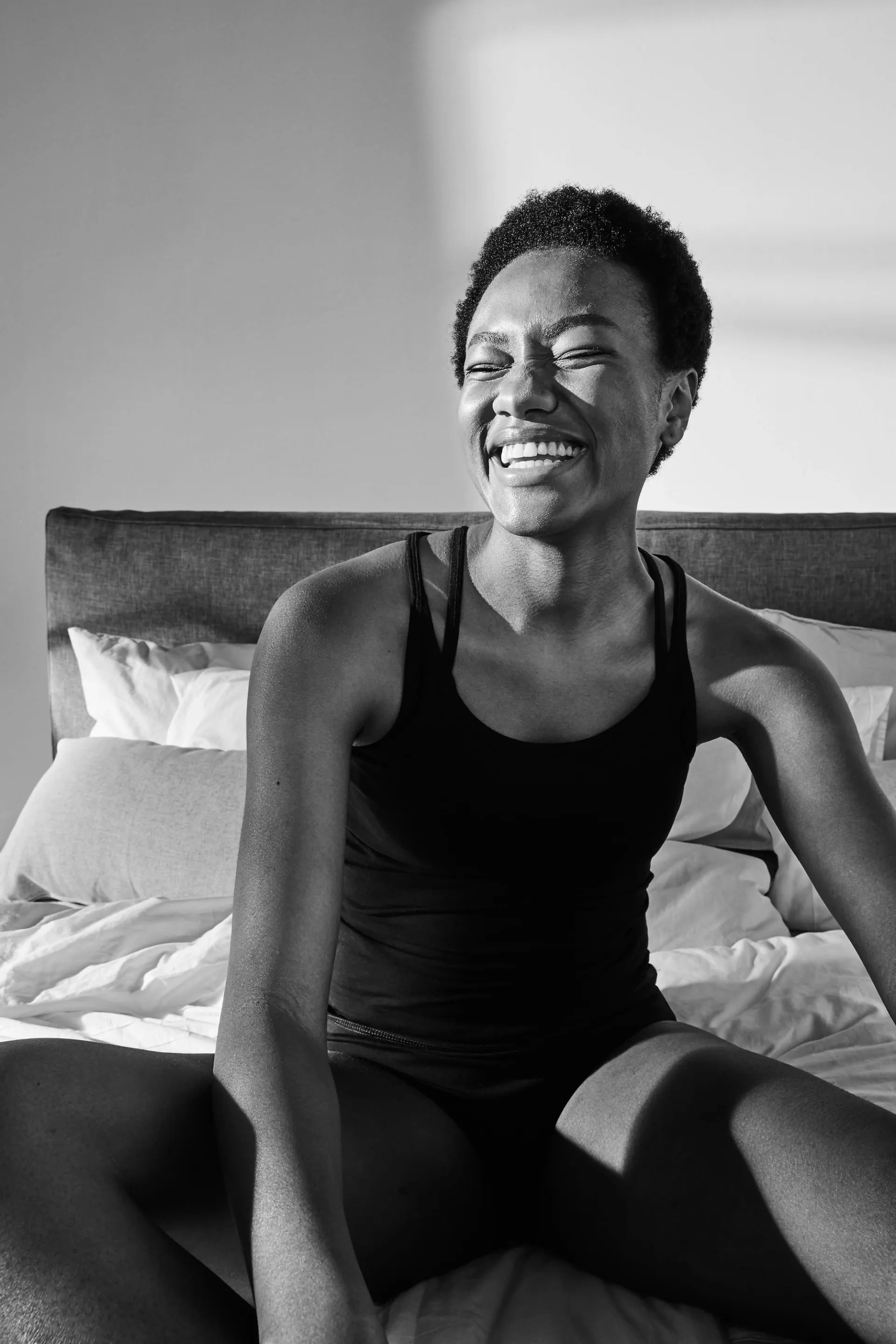 En person i svart linne och underkläder sitter avslappnat i en säng med vitt sänglinne. Solen kastar mjuka skuggor över kroppen och väggarna i rummet.