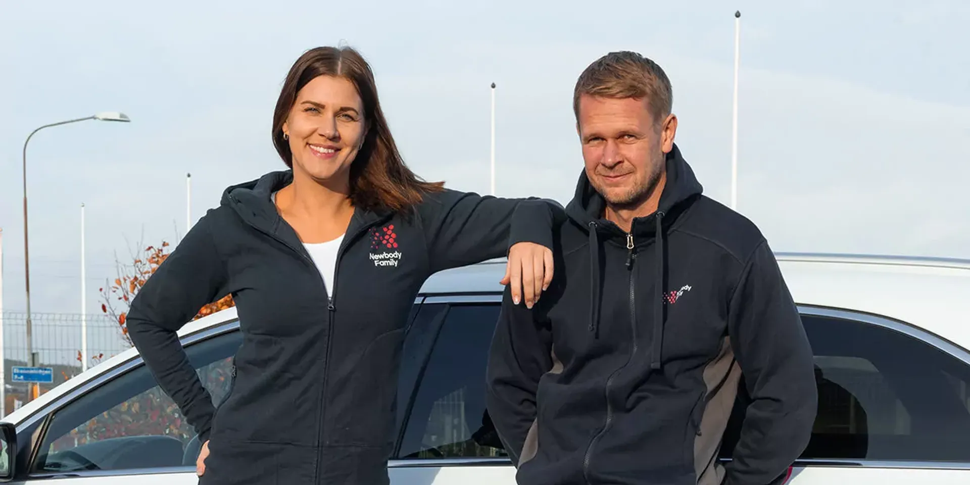 Två personer står lutade mot en vit bil, båda bär mörkblå hoodies med dragkedja och logotyp på bröstet. En kvinna och en man ser avslappnade och leende ut vid en utomhusmiljö.