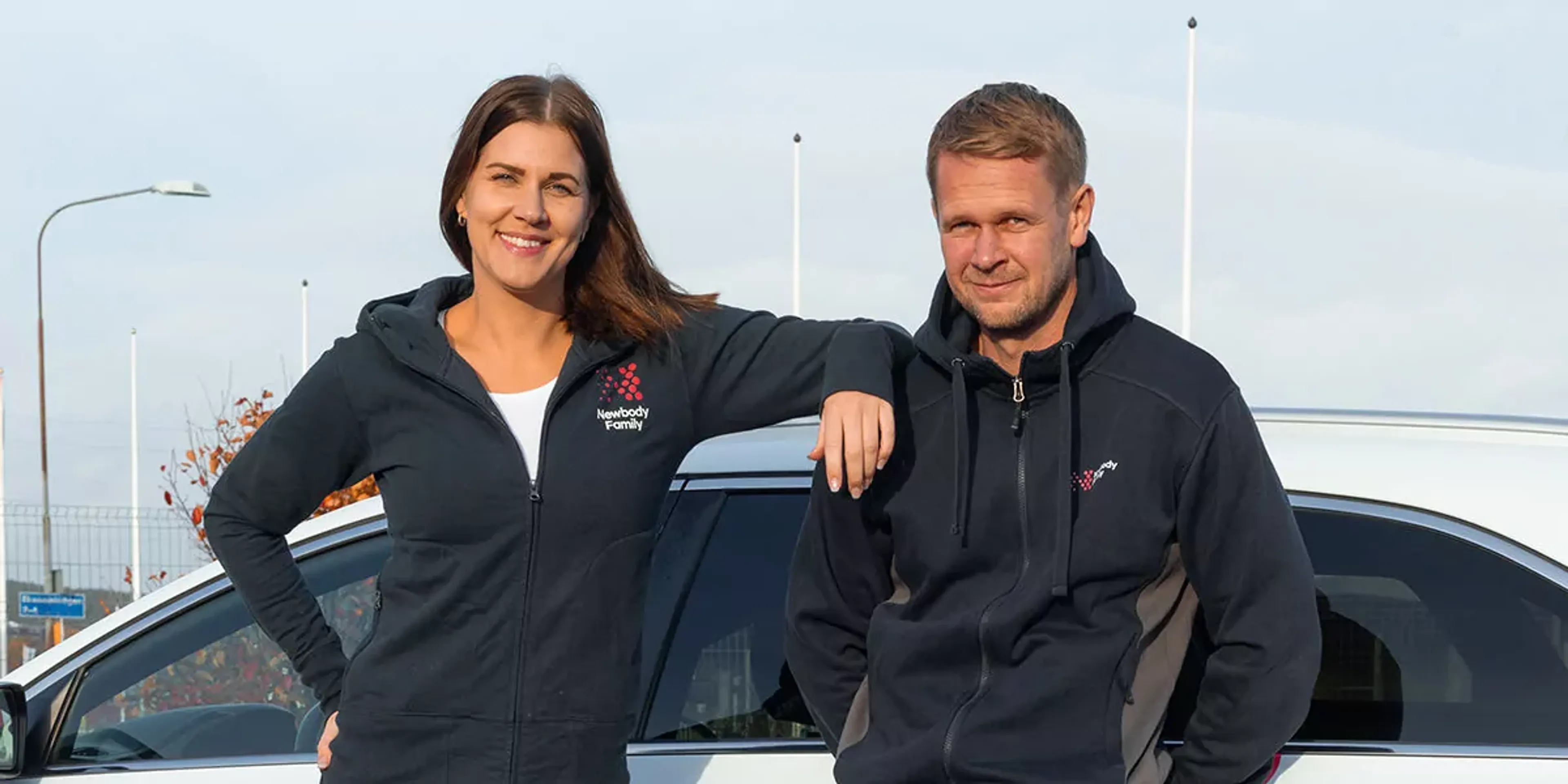 Två personer står lutade mot en vit bil, båda bär mörkblå hoodies med dragkedja och logotyp på bröstet. En kvinna och en man ser avslappnade och leende ut vid en utomhusmiljö.