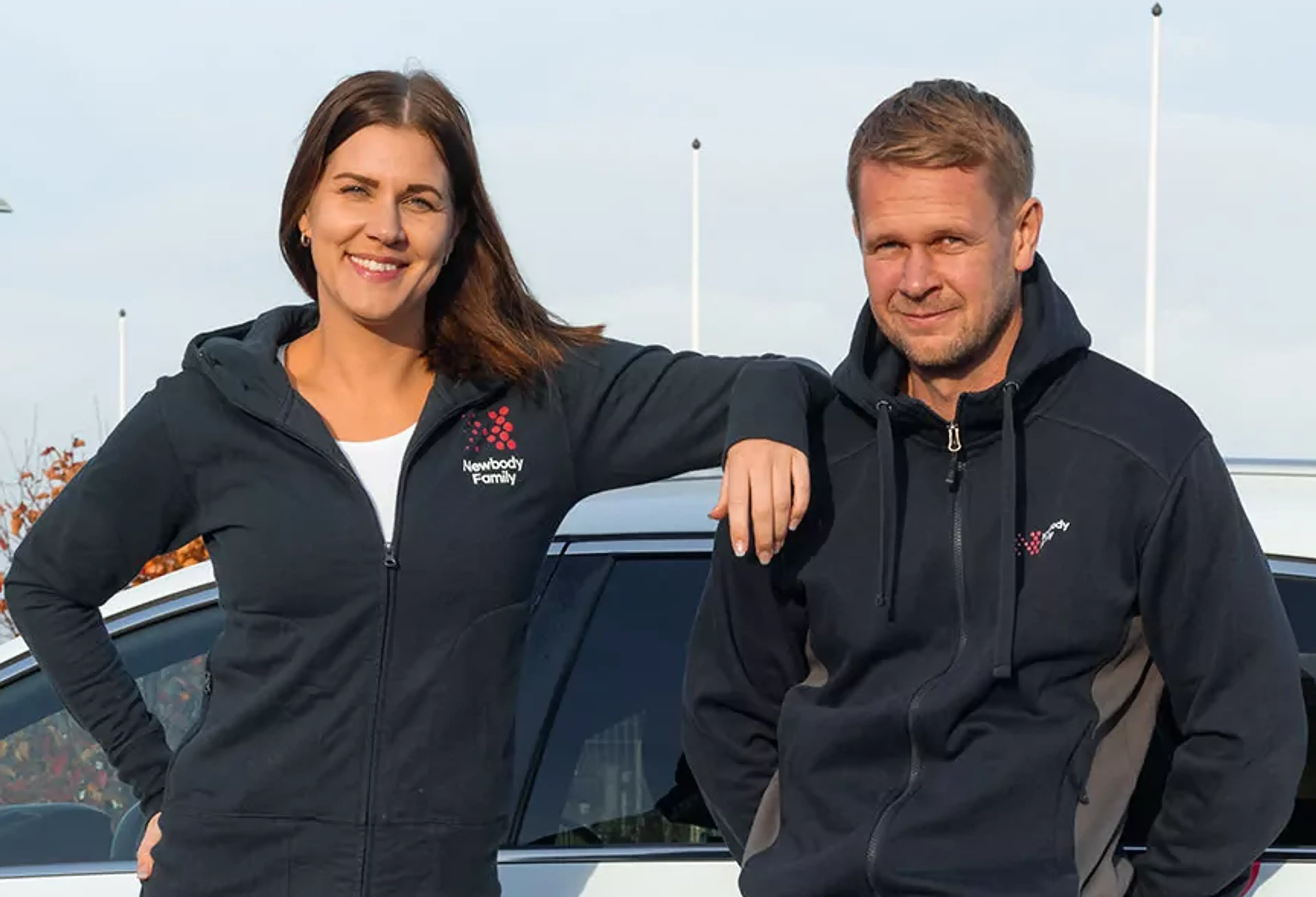 Två personer står lutade mot en vit bil, båda bär mörkblå hoodies med dragkedja och logotyp på bröstet. En kvinna och en man ser avslappnade och leende ut vid en utomhusmiljö.