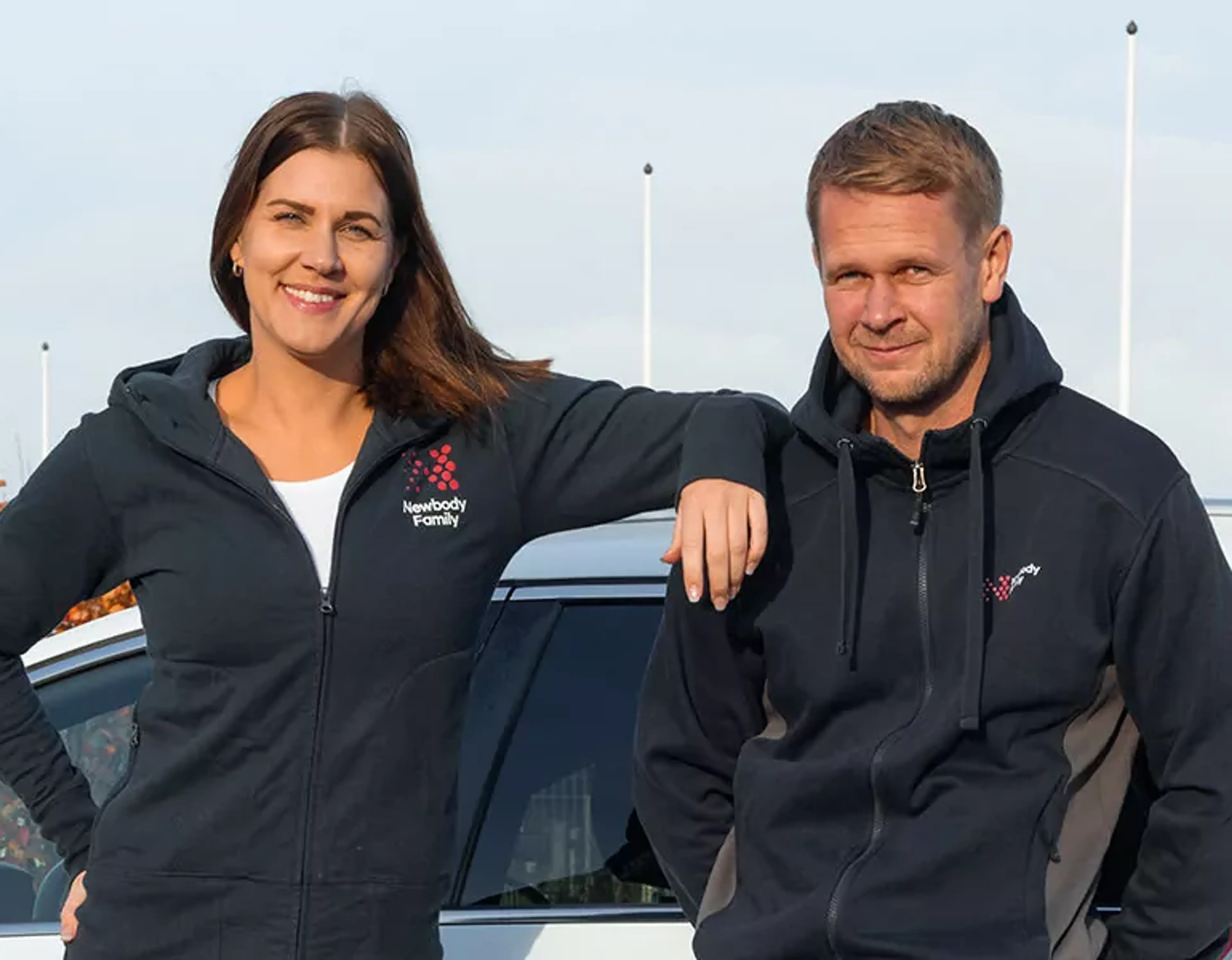 Två personer står lutade mot en vit bil, båda bär mörkblå hoodies med dragkedja och logotyp på bröstet. En kvinna och en man ser avslappnade och leende ut vid en utomhusmiljö.