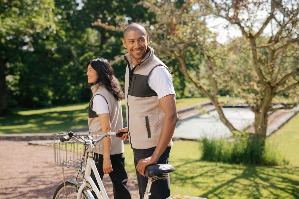 Två personer i mjuka, beige västar med svarta detaljer, båda bär vita t-shirts under och står utomhus en solig dag, en av dem håller en cykel med korg. Grönskande träd och en vattenyta syns i bakgrunden