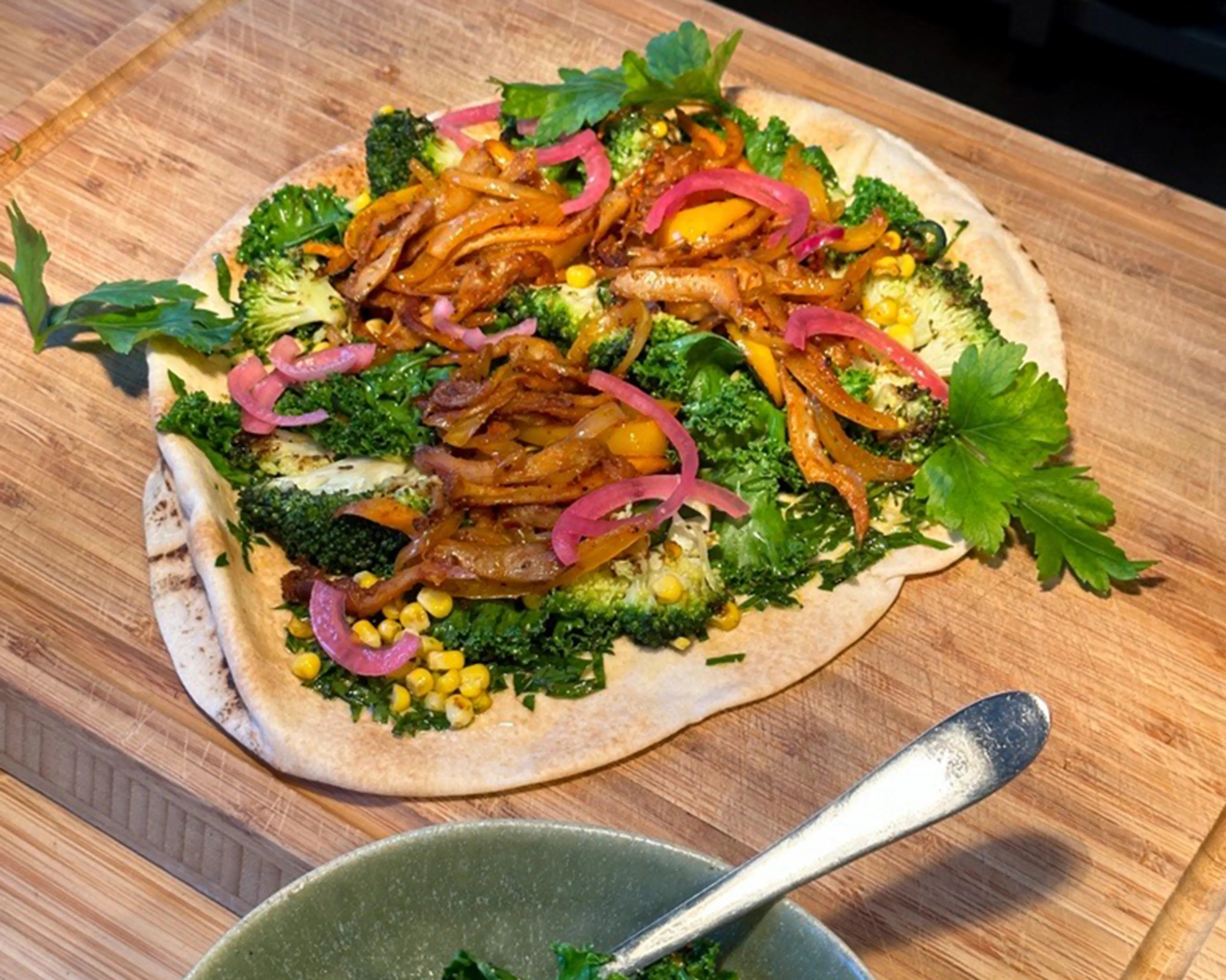 En tunn brödbotten toppad med stekt lök, broccoli, majs, grönkål, picklad rödlök och persilja. En fräsch och färgglad no-bake pizza på en skärbräda.