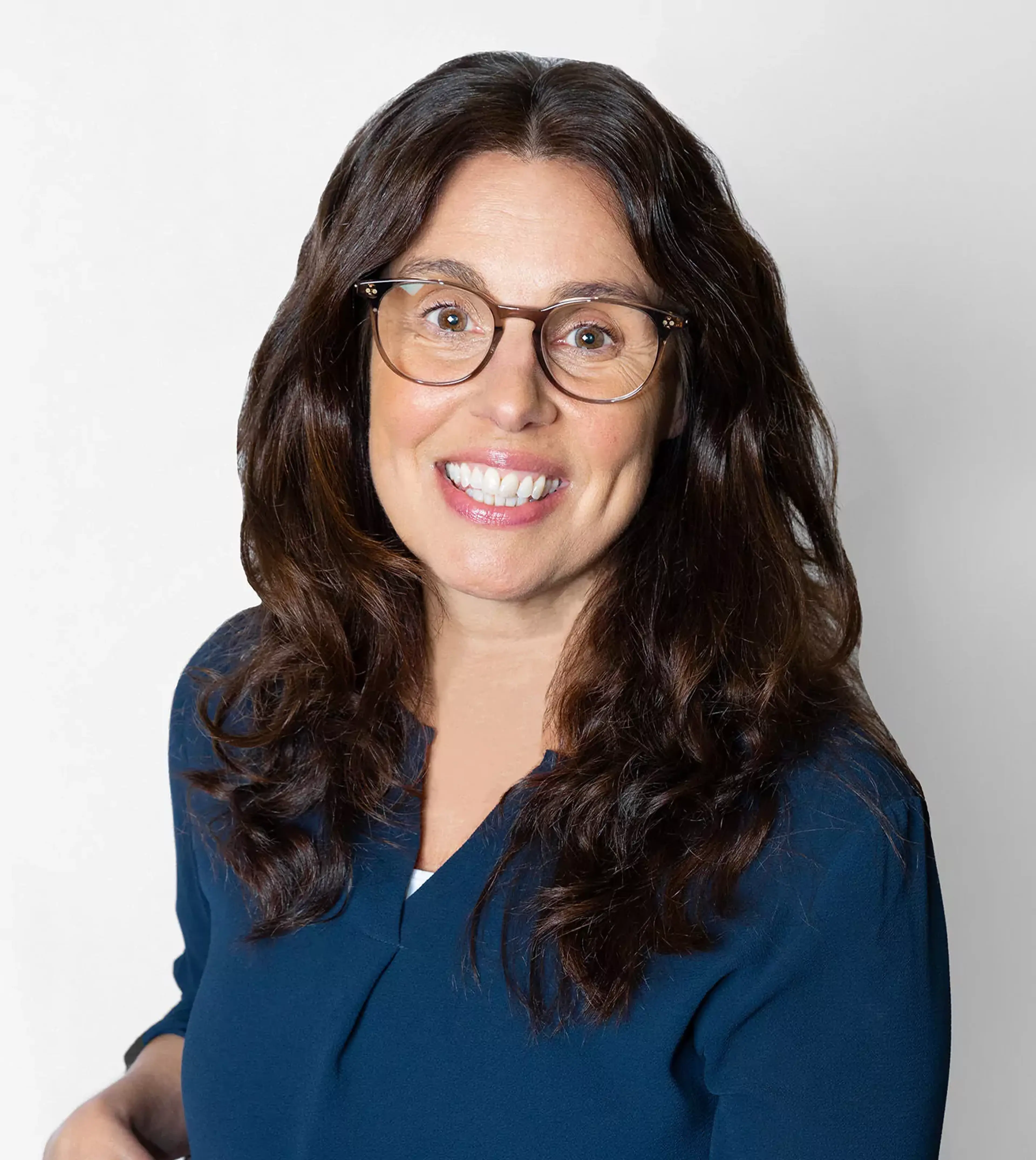 Kvinna med långt, mörkt hår och glasögon bär en mörkblå topp. Hon ler varmt mot kameran mot en ljus bakgrund.