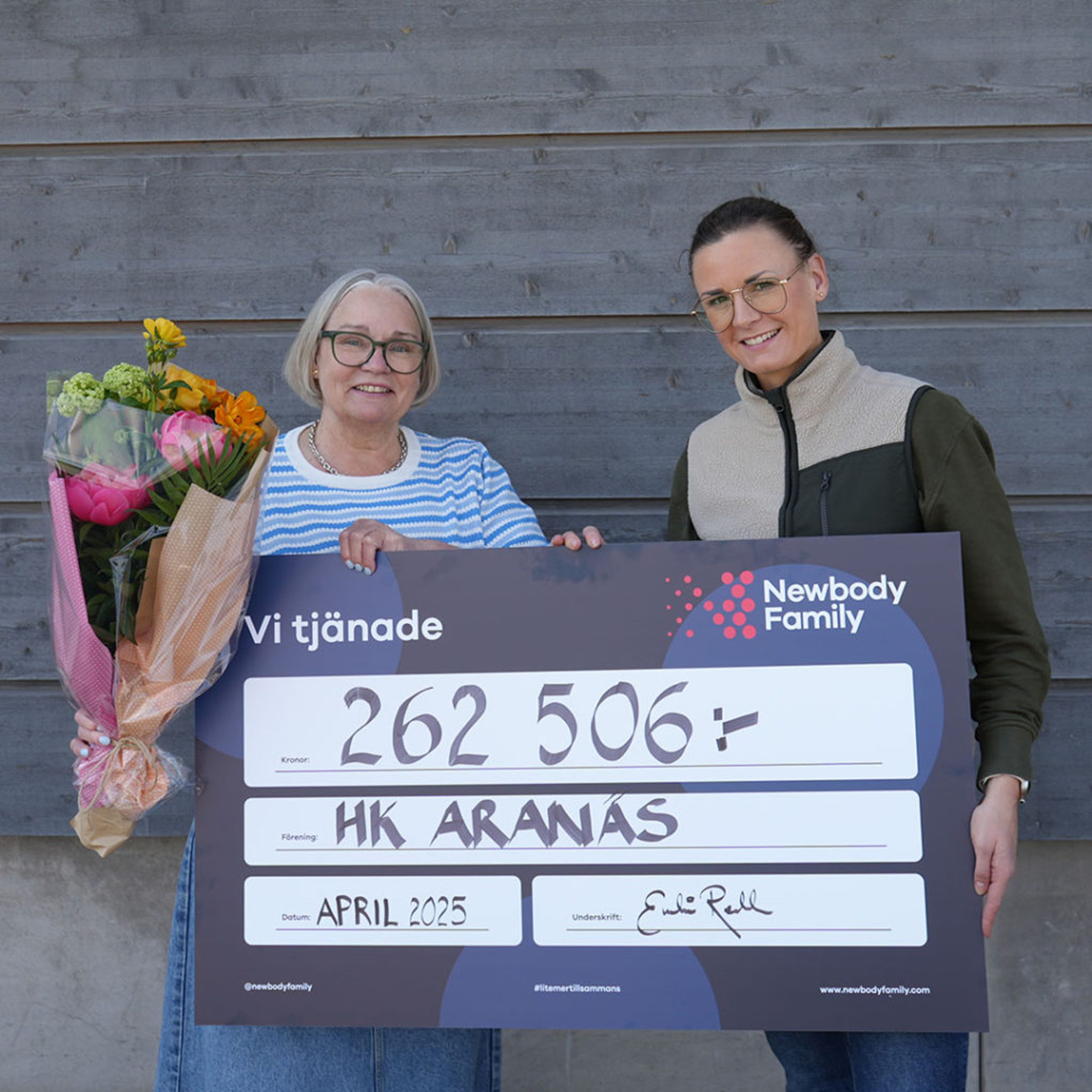 Två personer håller upp en stor check på 262 506 kronor till föreningen HK Aranäs, en av dem med en färgglad blombukett. Checken är daterad april 2025 och visar samarbetet med Newbody Family.
