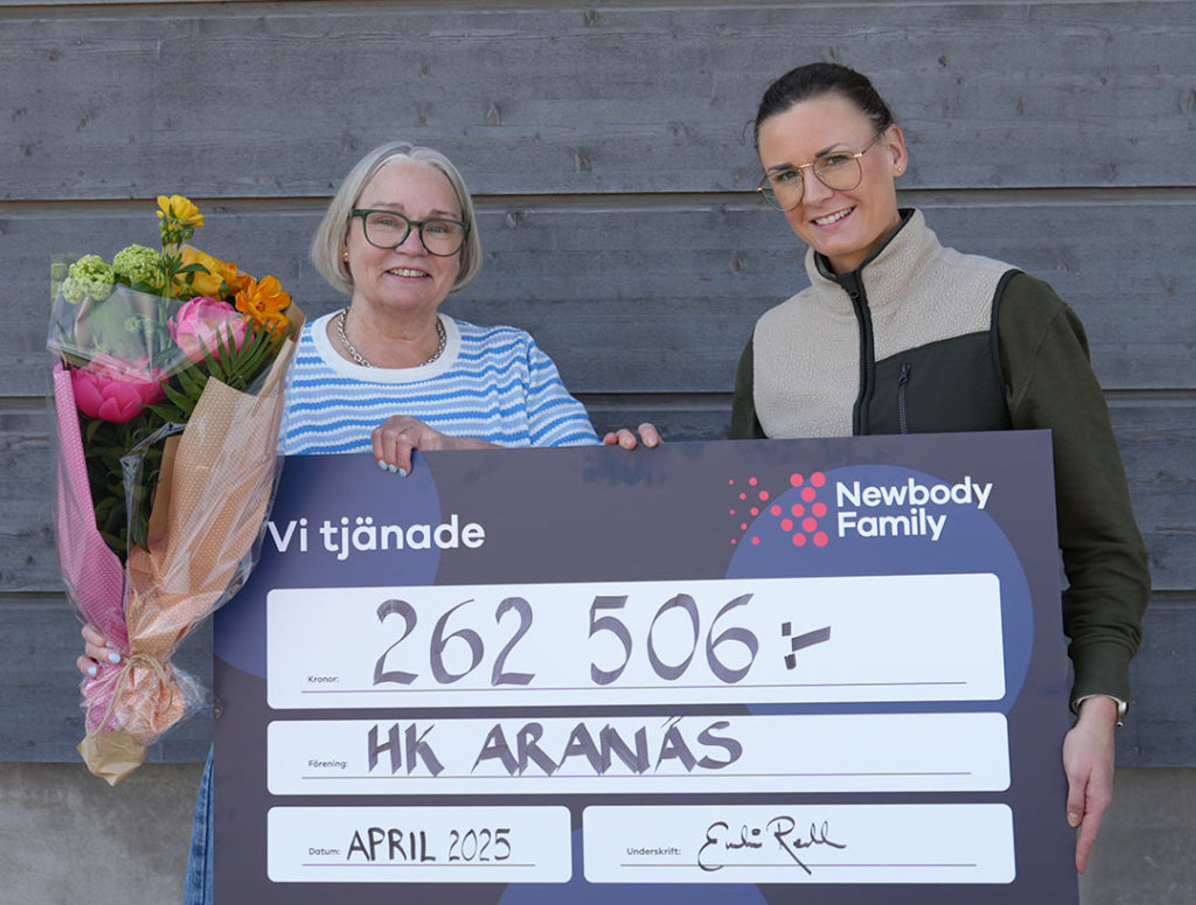 Två personer håller upp en stor check på 262 506 kronor till föreningen HK Aranäs, en av dem med en färgglad blombukett. Checken är daterad april 2025 och visar samarbetet med Newbody Family.
