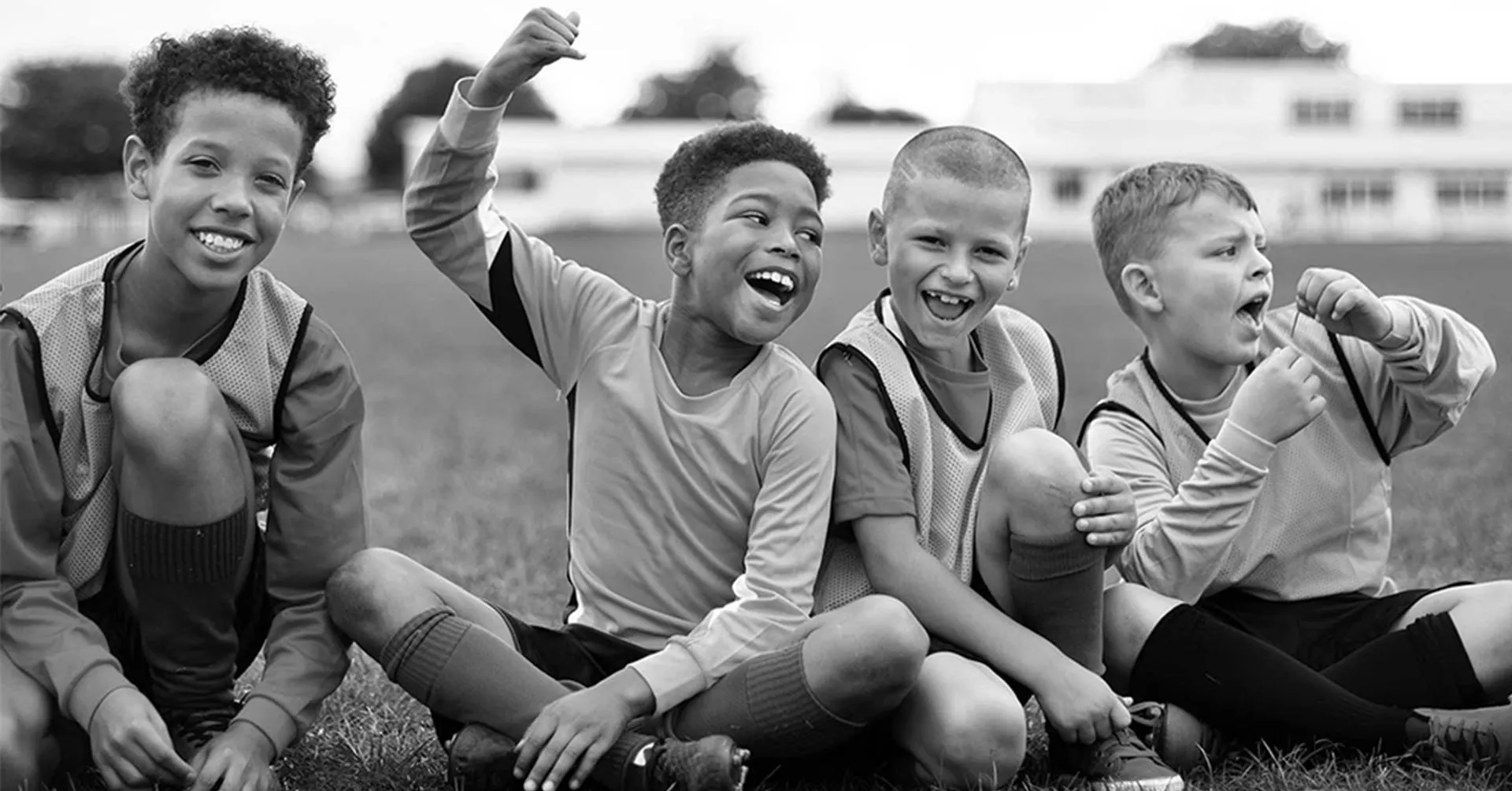 Fyra barn sitter på gräset i fotbollskläder och skrattar tillsammans efter en match eller träning. De visar glädje och samhörighet i en lekfull stund utomhus.