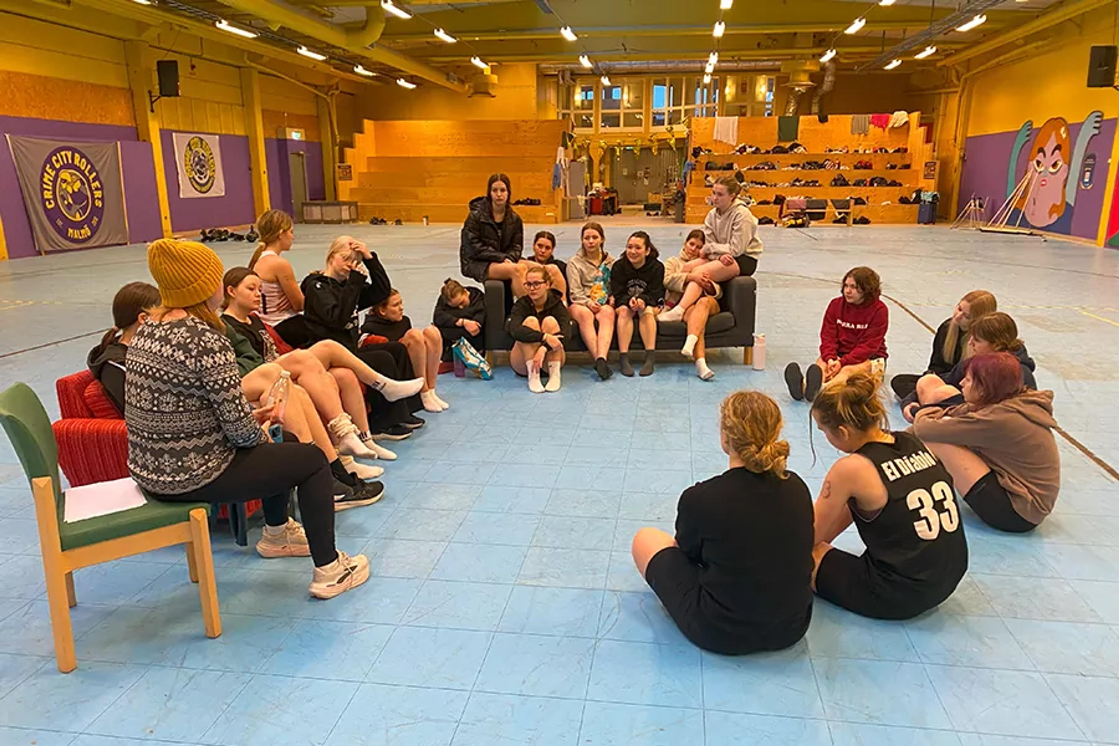 En grupp unga roller derby-spelare sitter i en cirkel på golvet och soffor i en stor hall med blått golv och gula väggar. Träningskläder och sportutrustning syns i bakgrunden.