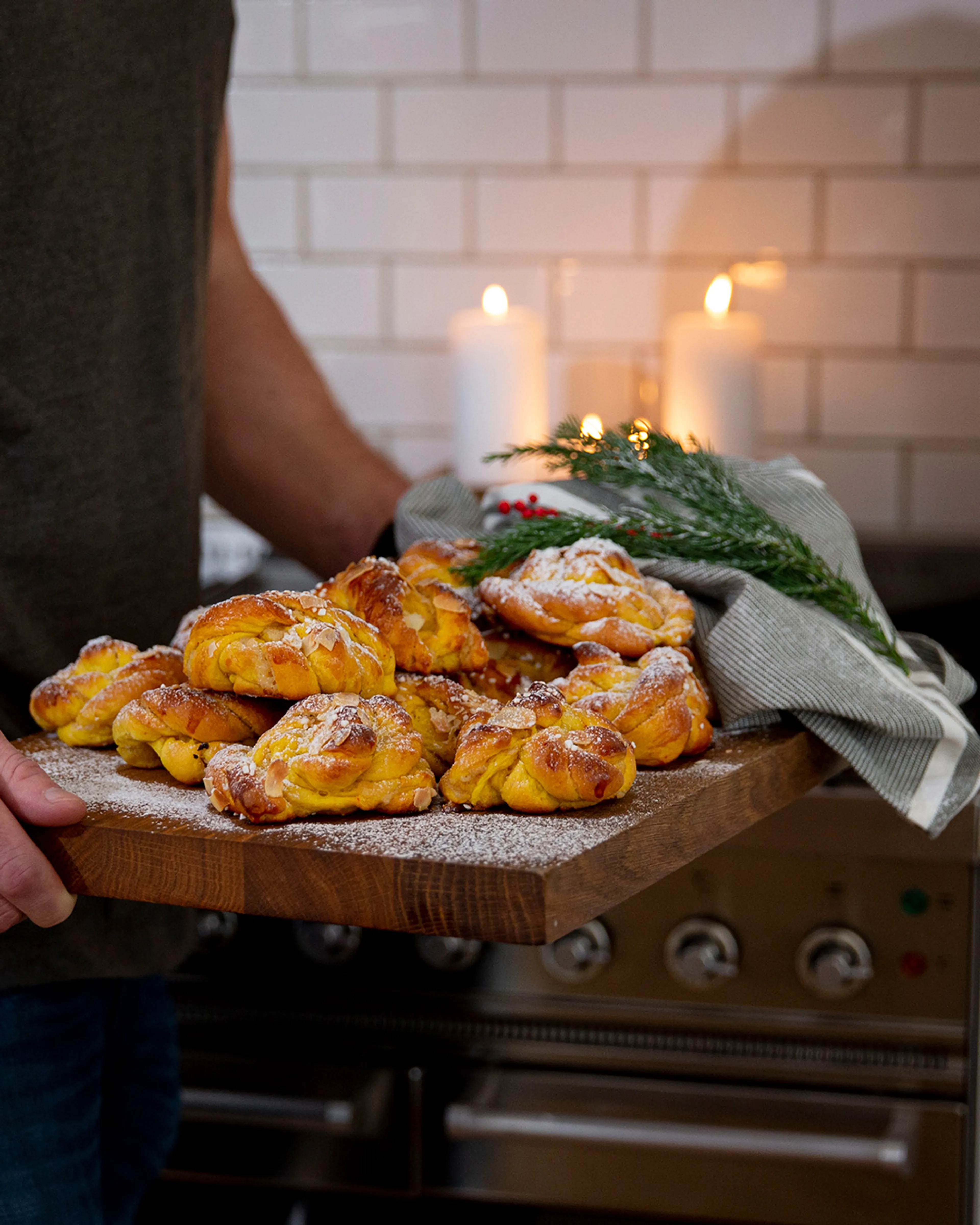 Nygräddade lussebullar på en träbricka, pudrade med florsocker och garnerade med mandelspån. En grön kvist med röda bär och två tända vita ljus i bakgrunden.