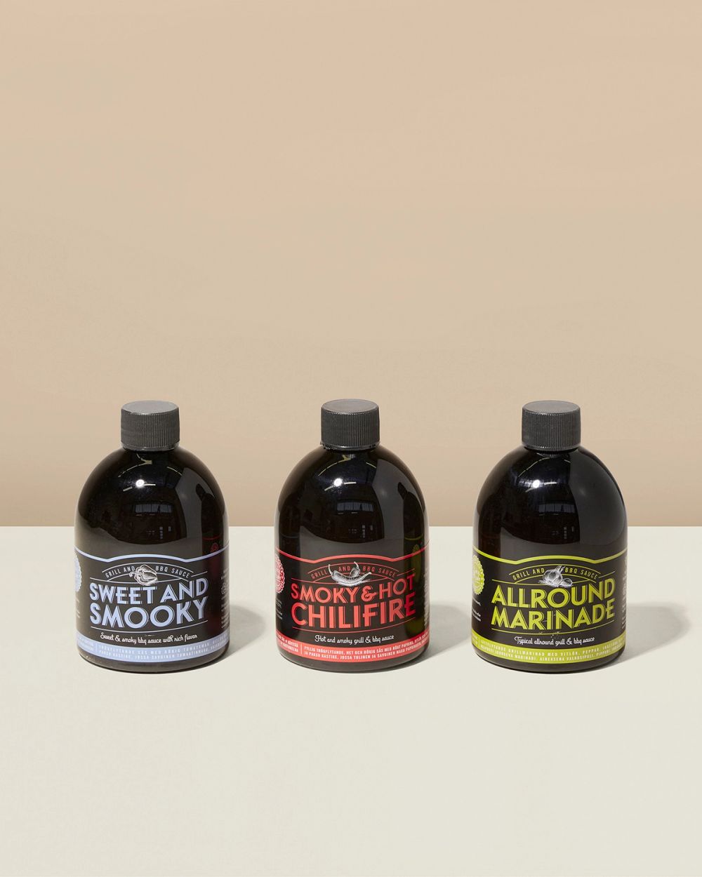 Tre mörka plastflaskor med olika grill- och BBQ-såser märkta i blått, rött och gult. Flaskorna står på en ljus yta mot en beige bakgrund.