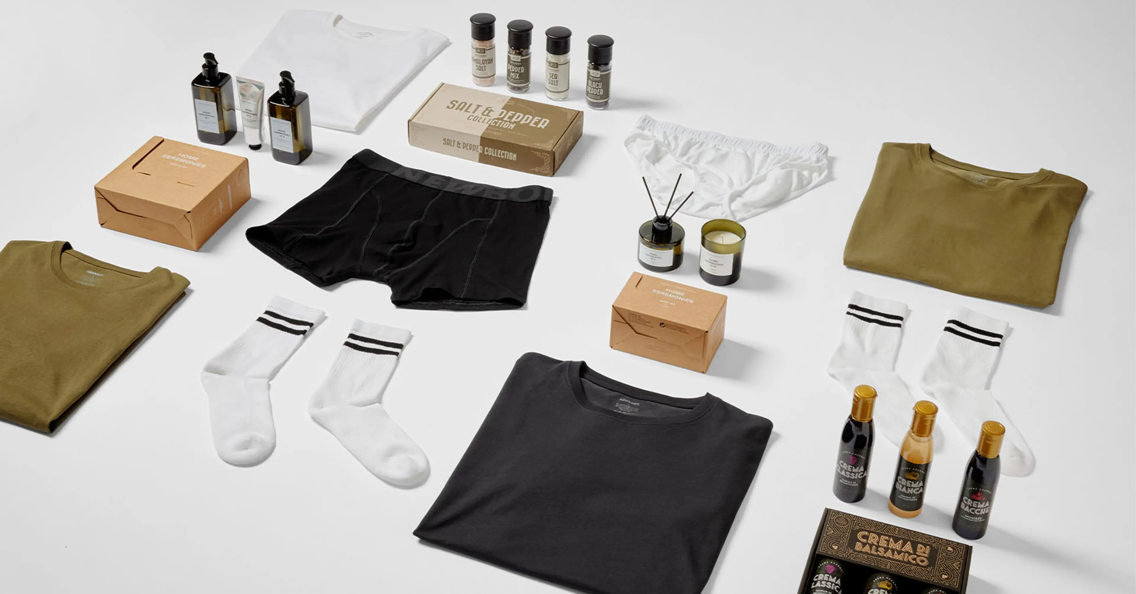 Uppsättningar av basplagg som t-shirts, strumpor och underkläder i neutrala färger, kompletterade med lyxiga doftljus, kryddor och balsamicosåser. Produkterna ligger prydligt arrangerade mot en beige bakgrund