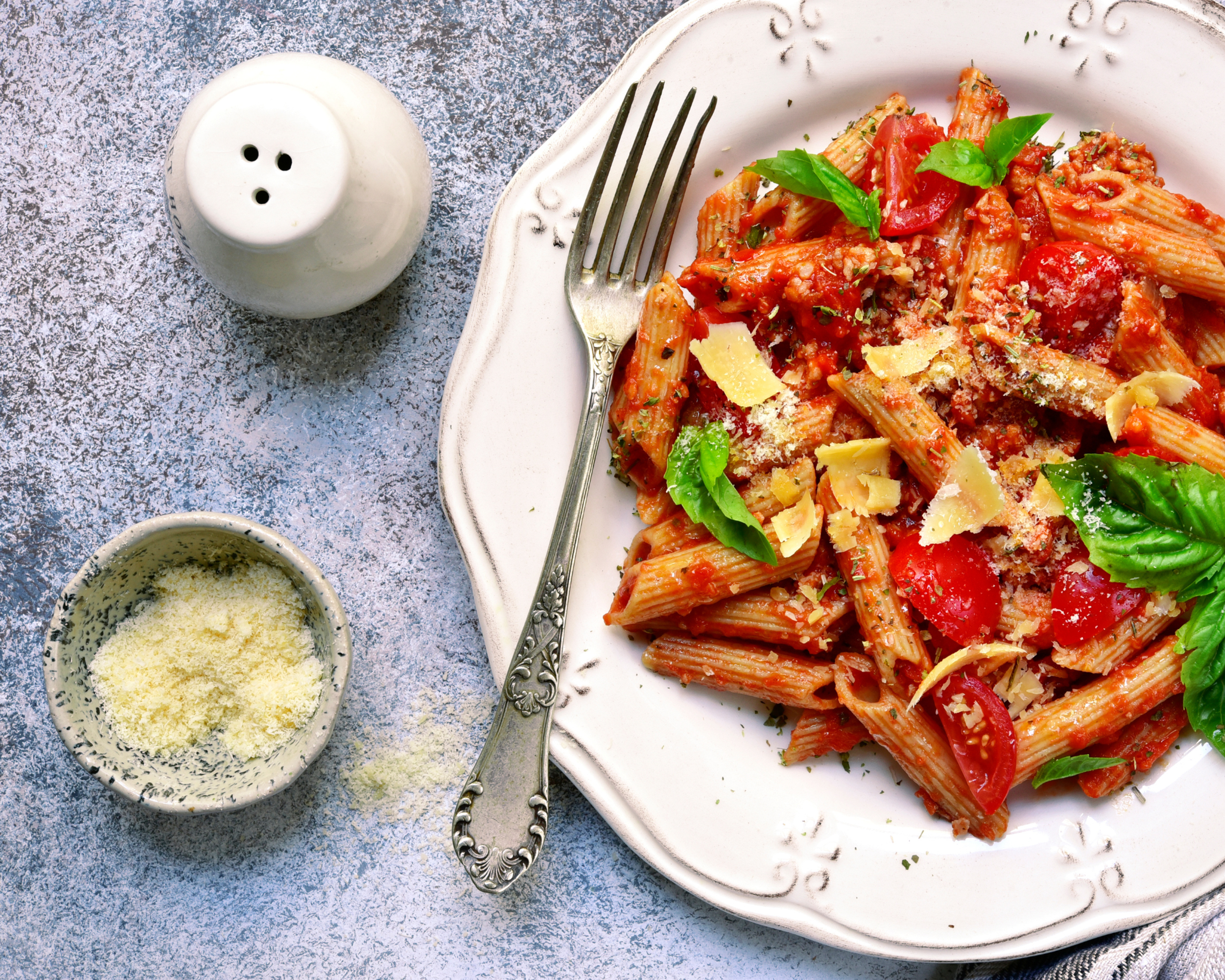 En tallrik med pennepasta blandad med tomatsås, körsbärstomater, basilika och riven ost. Bredvid ligger en liten skål med mer riven ost och en vit saltkar.