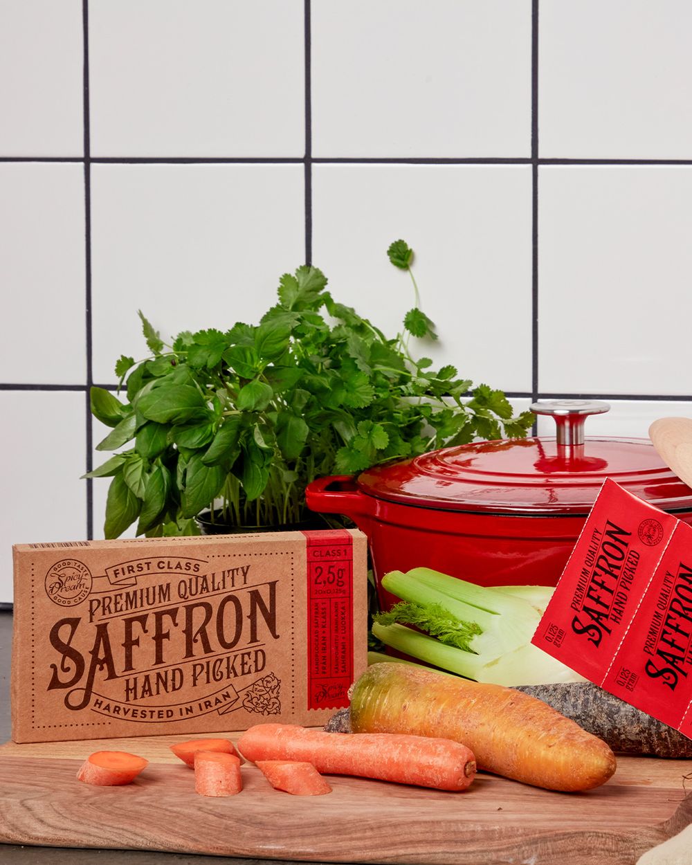 Ett paket saffran med handplockade trådar ligger på en skärbräda tillsammans med morötter, fänkål och färska gröna örter, framför en röd gjutjärnsgryta.
