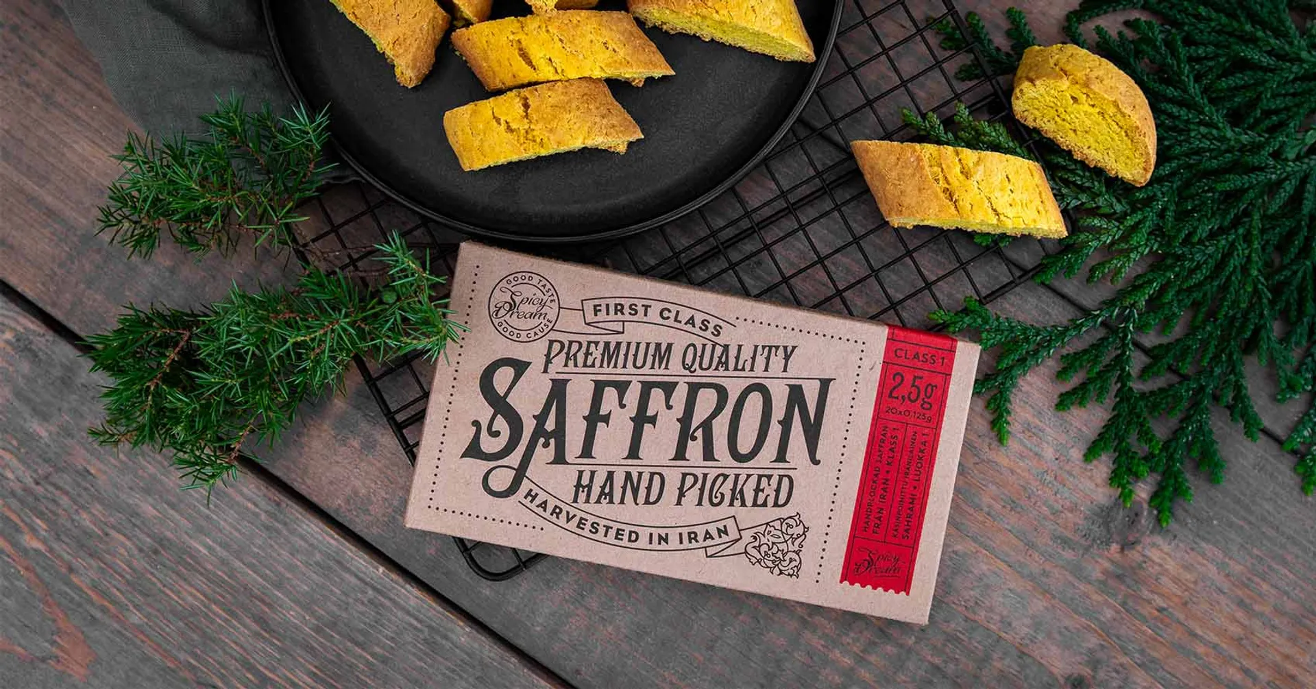 Ett paket med handplockad saffran från Iran ligger på ett bord bredvid flera bitar av gulbrunt bakverk på en svart plåt. Gröna kvistar pryder bilden för en naturlig känsla.