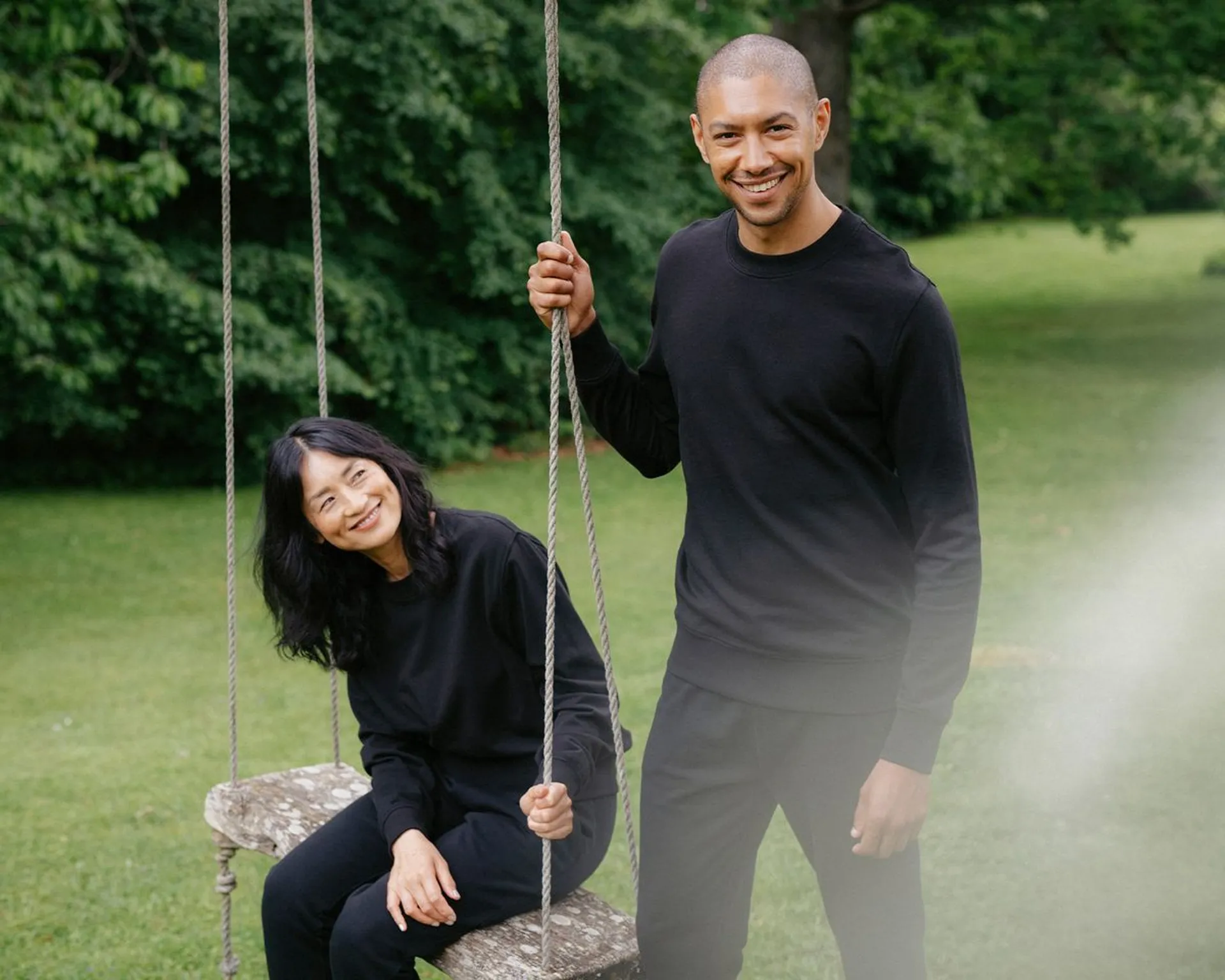 Två personer i svarta, bekväma kläder är utomhus vid en trägunga i grönskande park. Den ena sitter på gungan och ler medan den andra står bredvid och håller i repet.