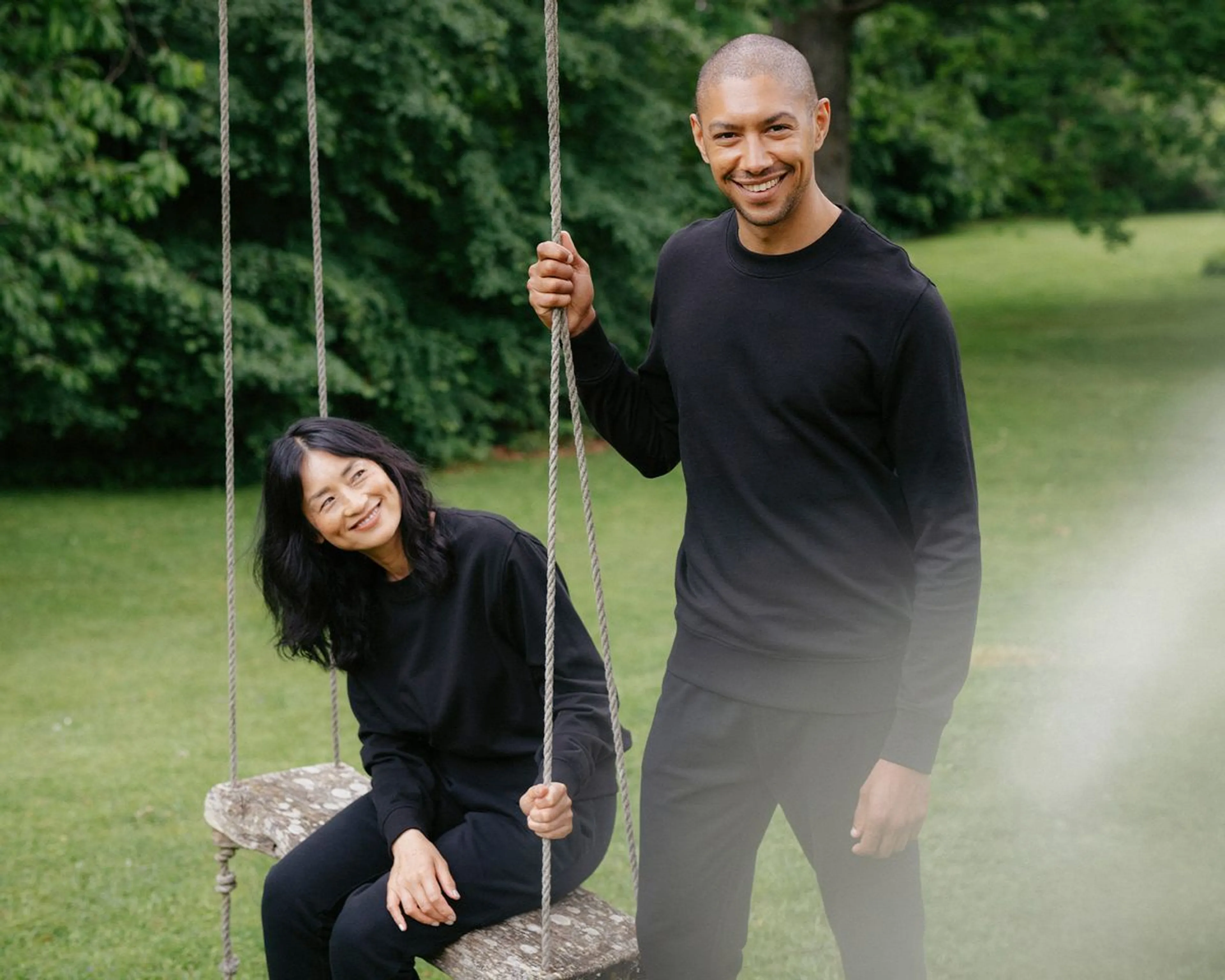 Två personer i svarta, bekväma kläder är utomhus vid en trägunga i grönskande park. Den ena sitter på gungan och ler medan den andra står bredvid och håller i repet.