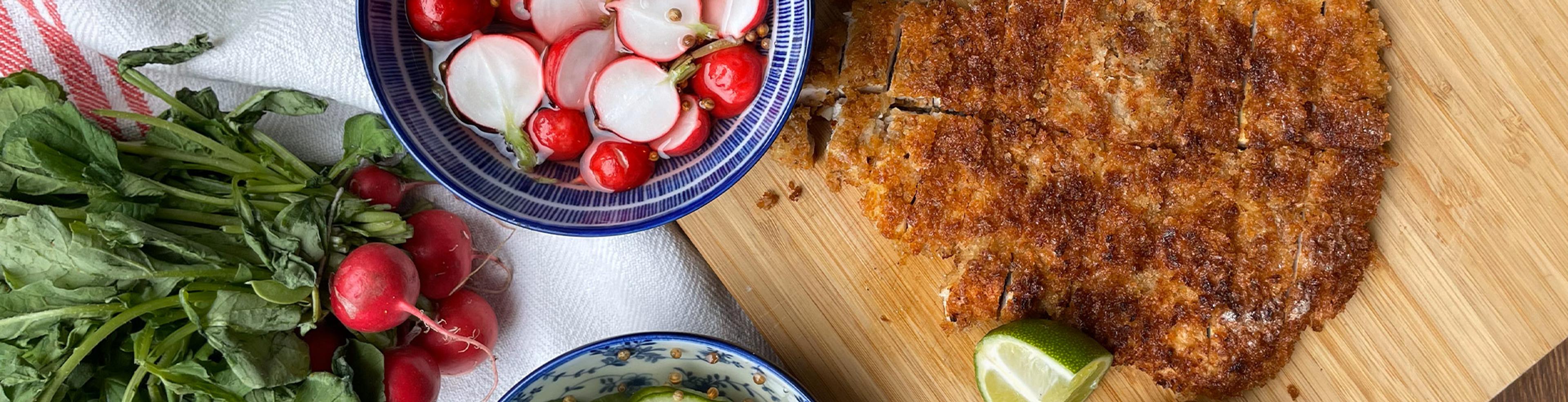 Krispigt panerad kyckling på en skärbräda med limeklyfta, bredvid en skål med skivade rädisor och en bunt färska rädisor.