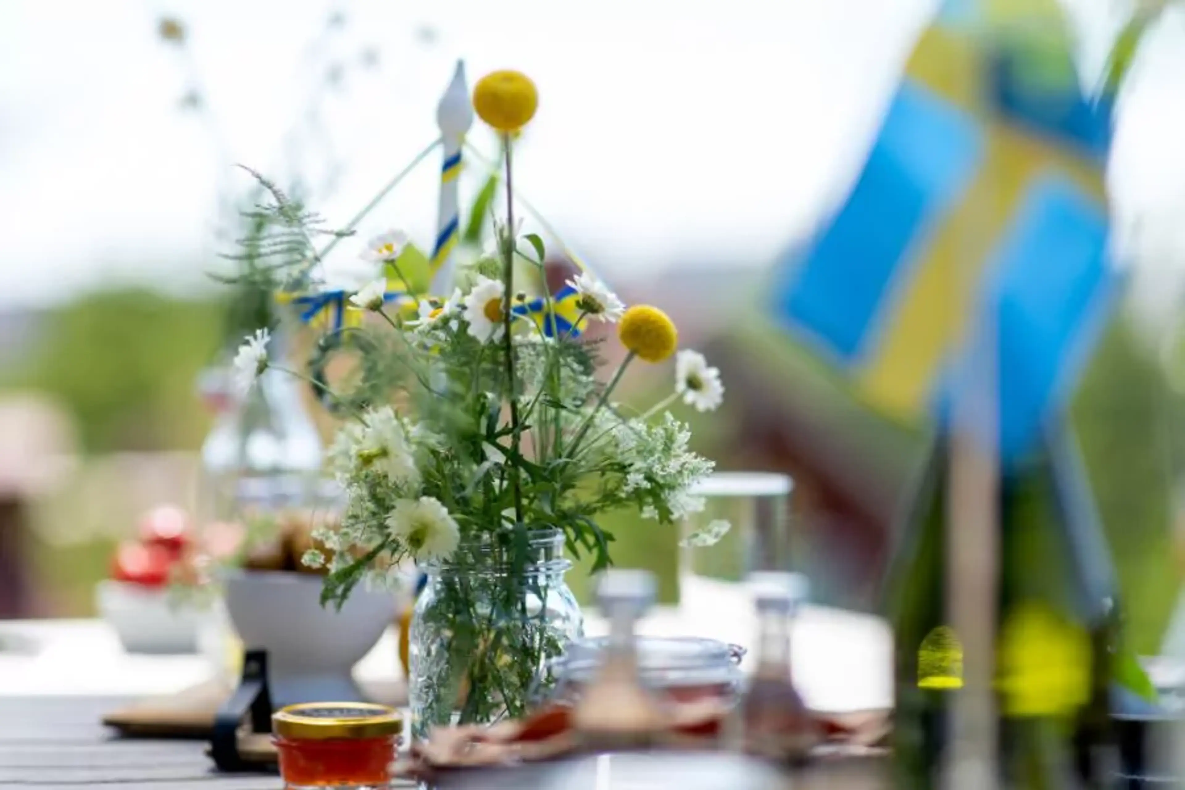 En vas med sommarblommor och små svenska flaggor står på ett bord med olika mat- och glasburkar runtom. Bakgrunden visar en grönskande, solig utemiljö.