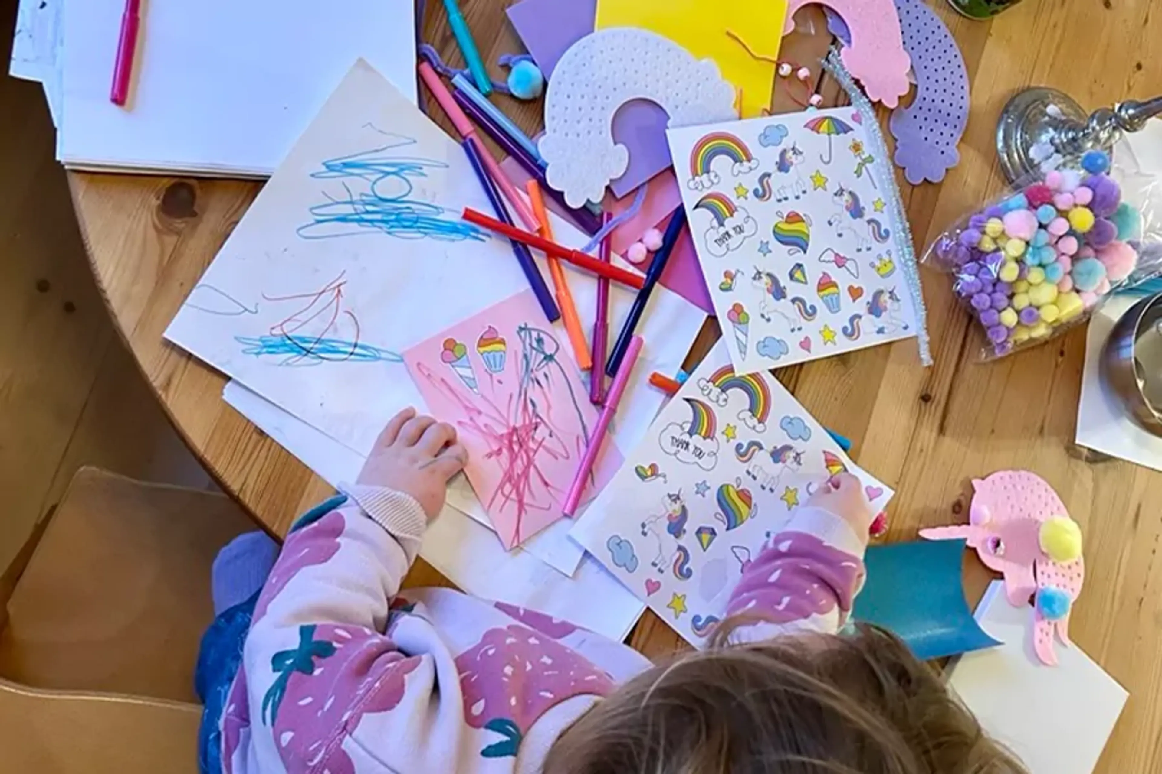 Barn sitter vid ett träbord fullt av färgglada pennor, ritpapper och klistermärken med regnbågar och enhörningar. En påse med små, mjuka bollar i pasteltoner ligger också på bordet.