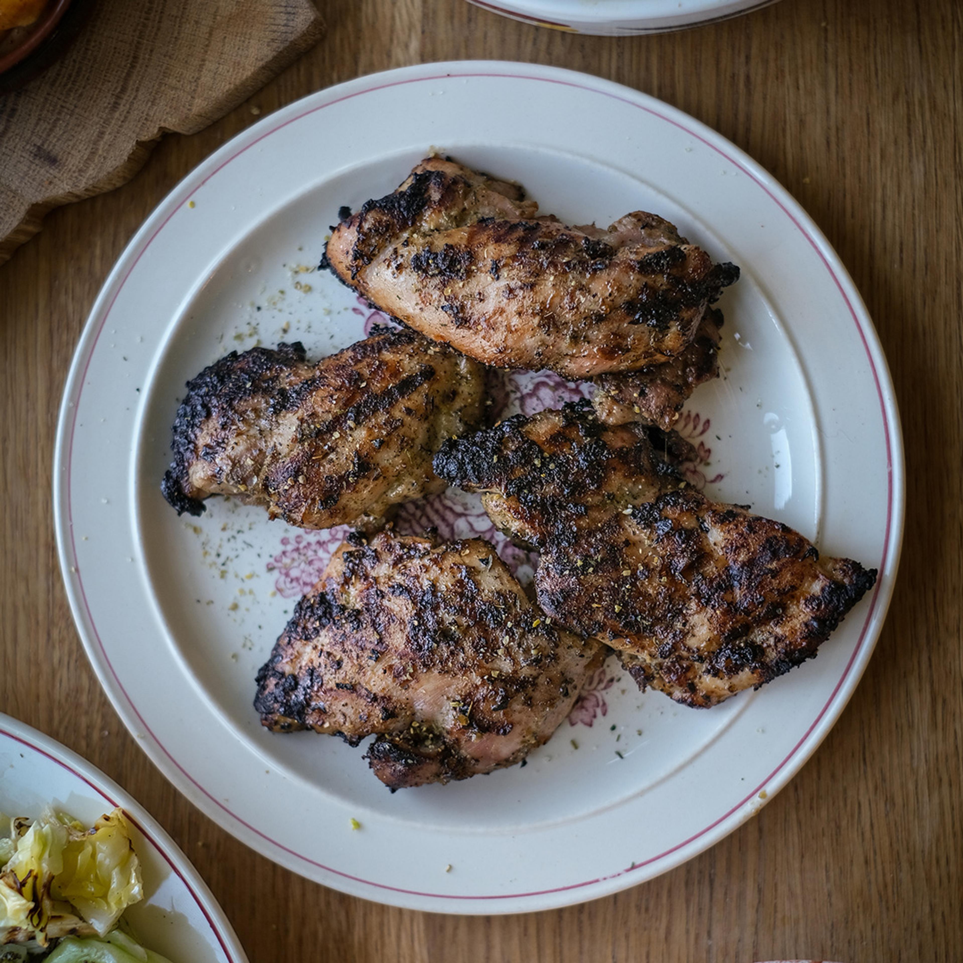 Fem grillade kycklinglårfiléer med en kryddig yta, serverade på en vit tallrik med rosa kant. Ytan är lätt kolgrillad med synliga grillränder.
