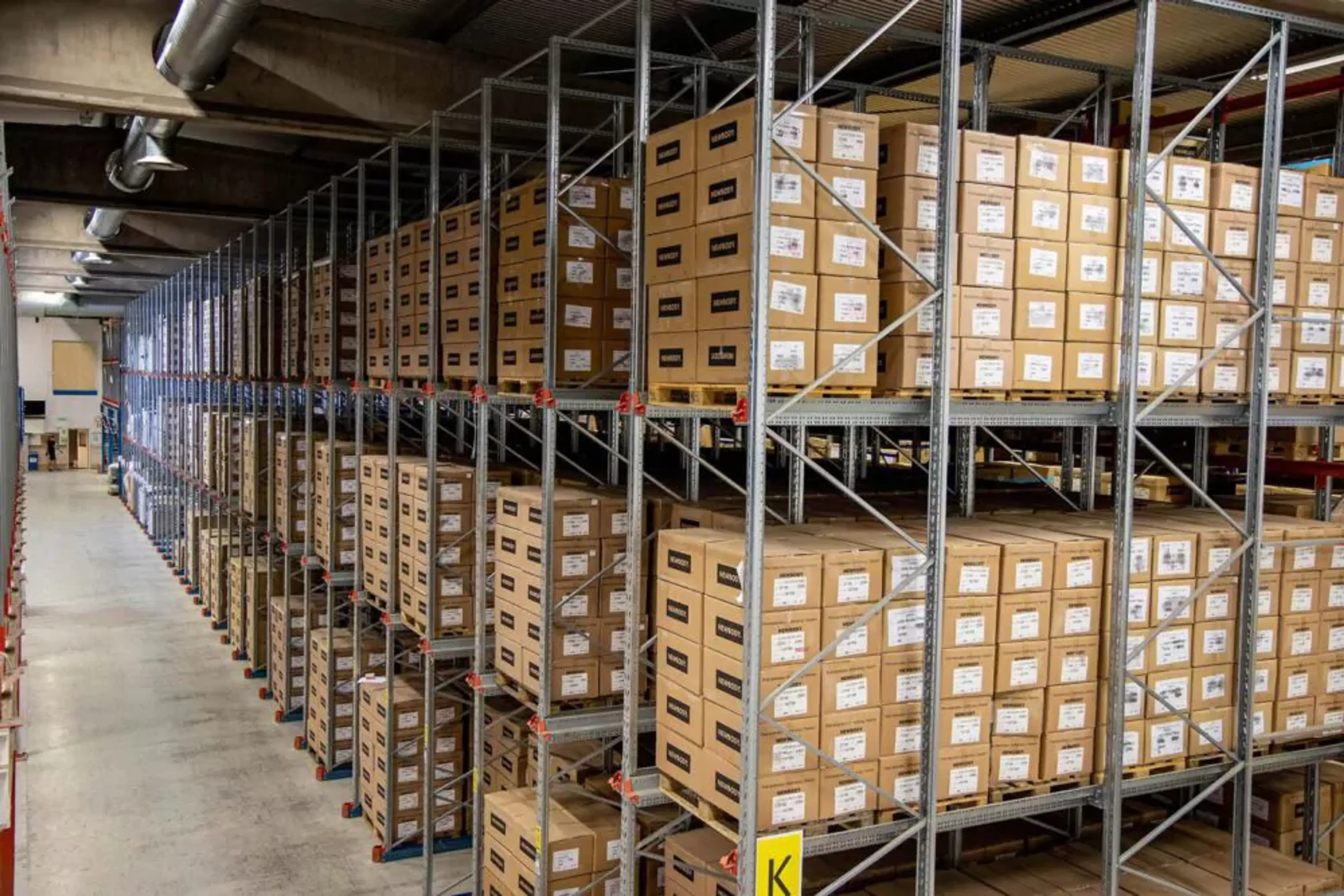 Hyllor fyllda med prydligt staplade kartonger i ett stort lagerutrymme. En översikt av välorganiserad förvaring i en ljus och rymlig lokal.