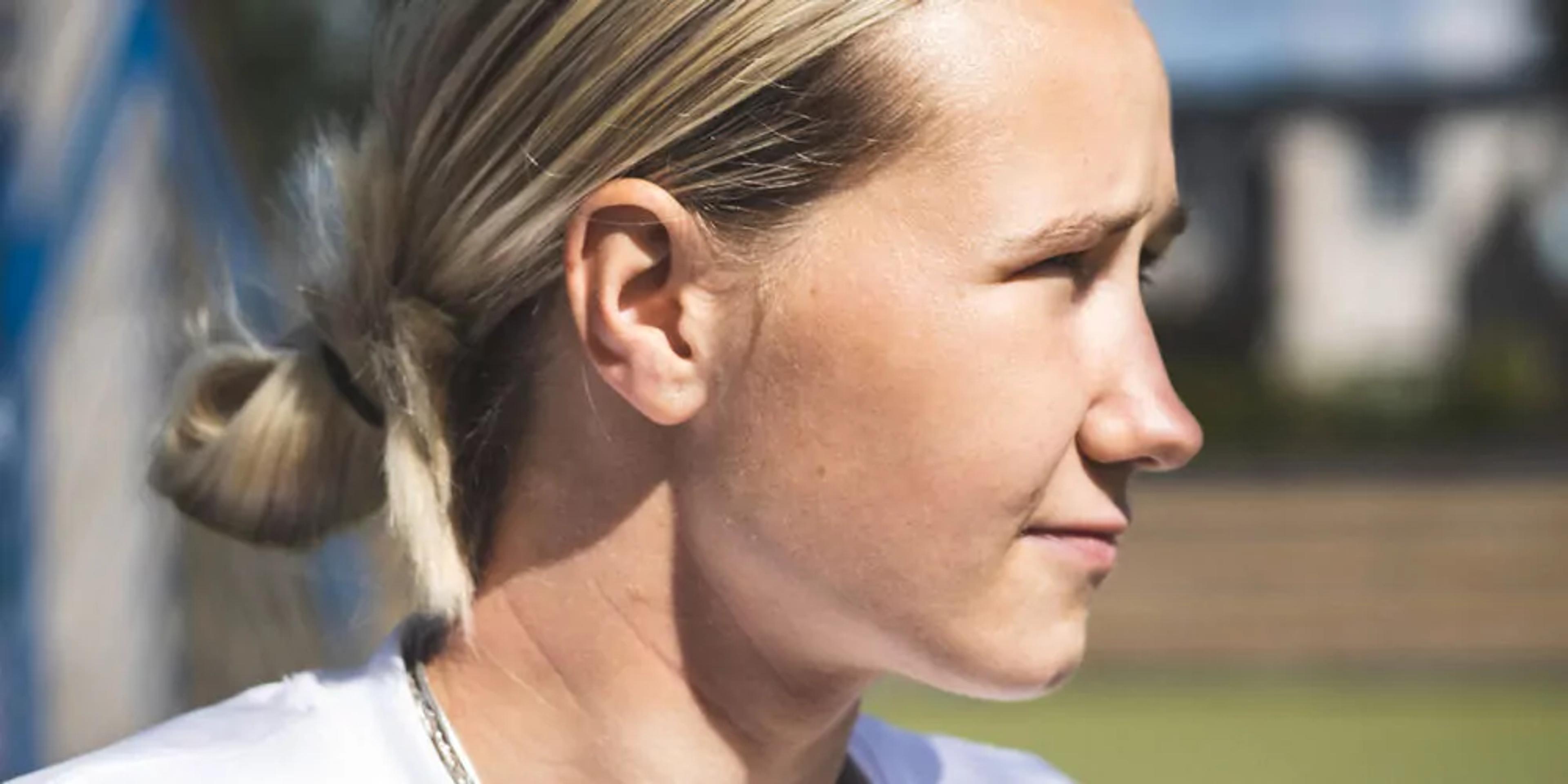 En person med blont hår uppsatt i en låg knut, iförd vit tröja, blickar mot något utanför bild. Bakgrunden är suddig med gröna och blå toner.