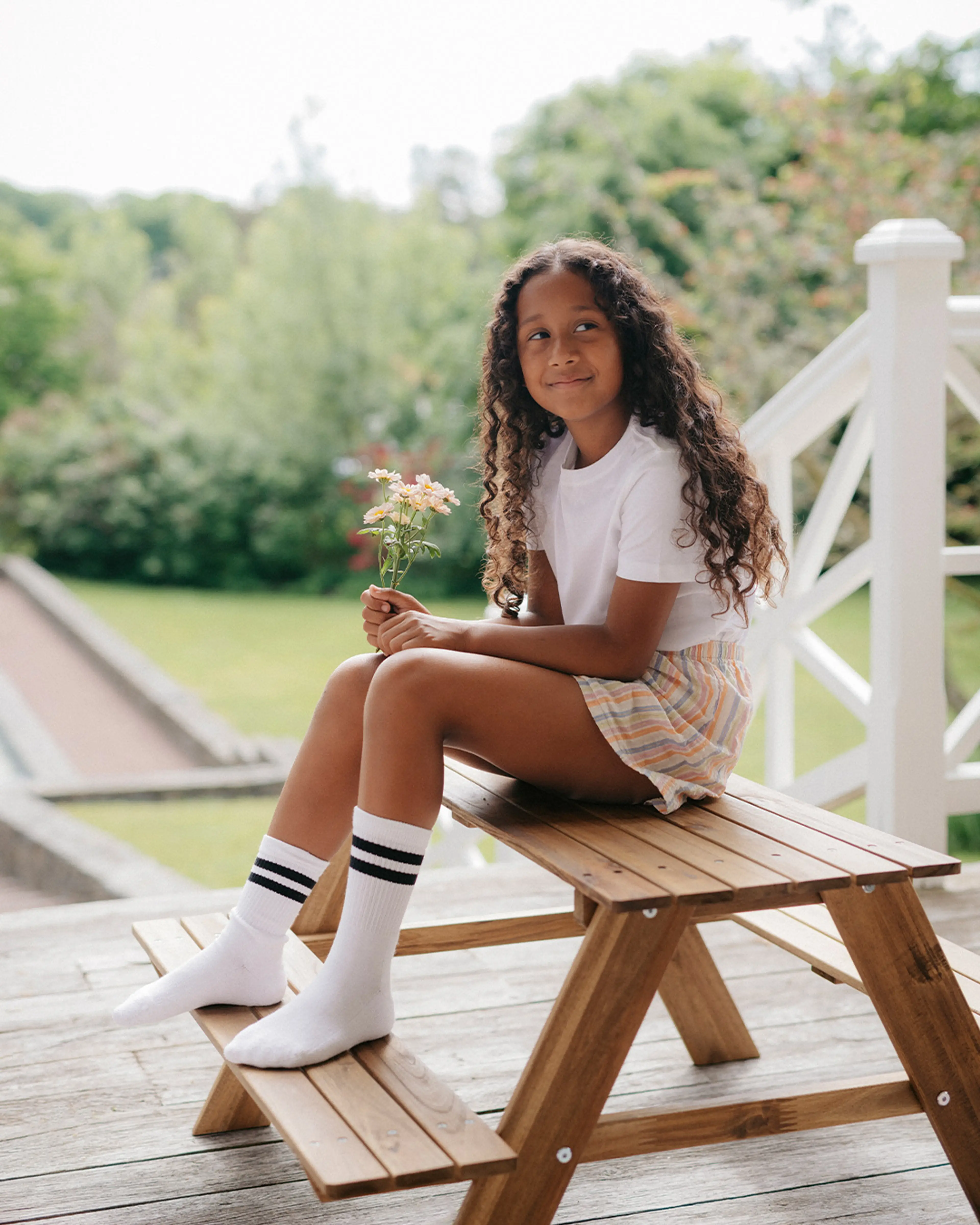 En ung person sitter på ett träpiknikbord utomhus, klädd i en vit t-shirt, randiga shorts och vita strumpor med svarta ränder, medan hen håller en liten bukett blommor. Bakgrunden visar grönskande träd och