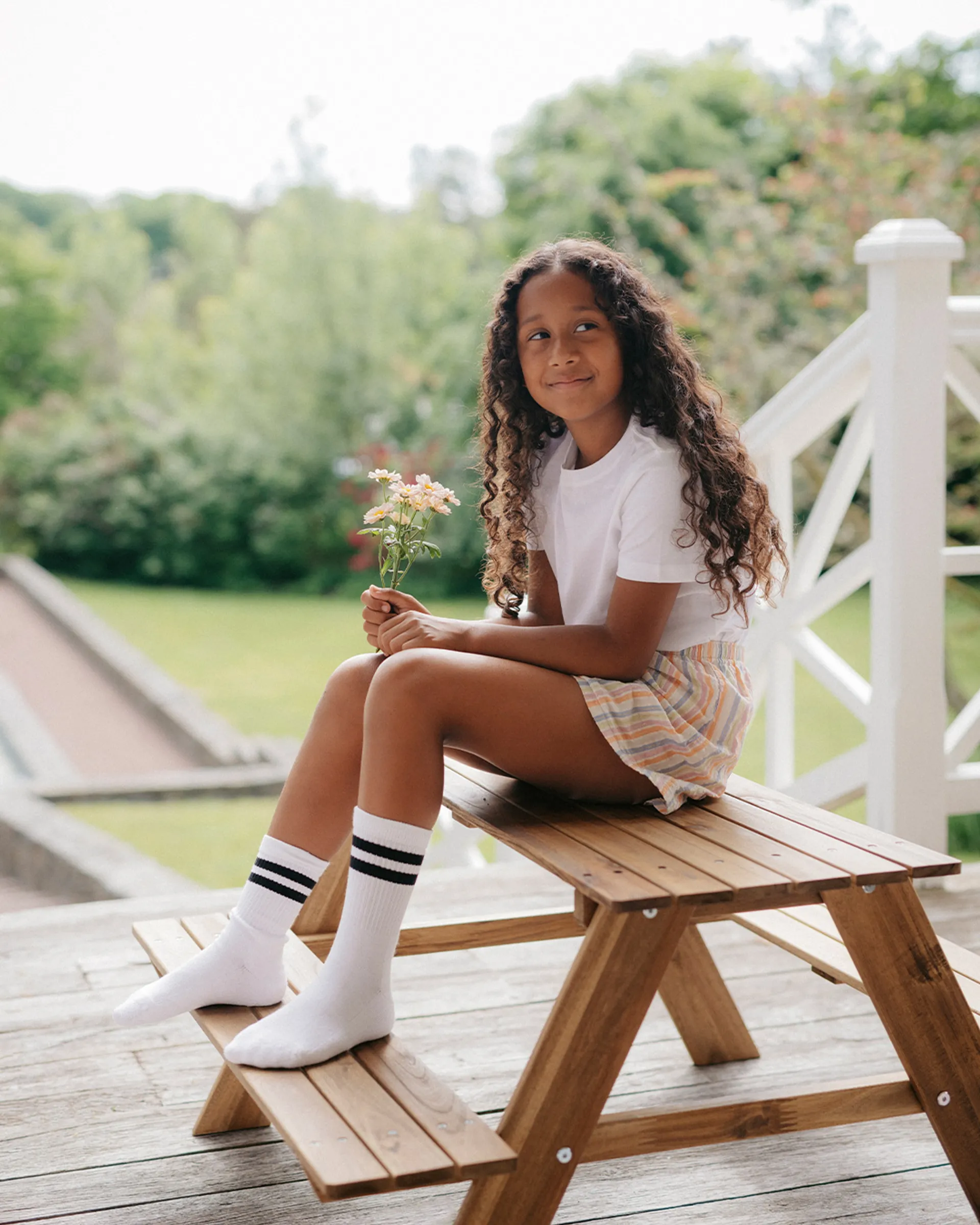 En ung person sitter på ett träpiknikbord utomhus, klädd i en vit t-shirt, randiga shorts och vita strumpor med svarta ränder, medan hen håller en liten bukett blommor. Bakgrunden visar grönskande träd och