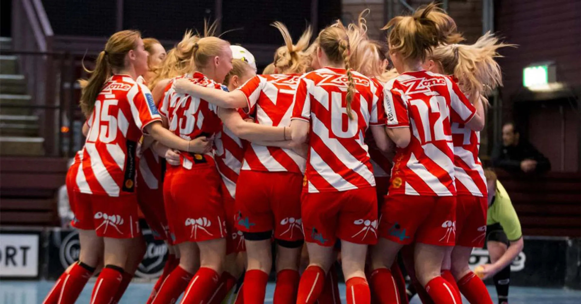 Ett lag med innebandytjejer i röda och vita matchkläder samlas i en grupp på en inomhusplan. De har nummer på ryggen och sportiga skor.