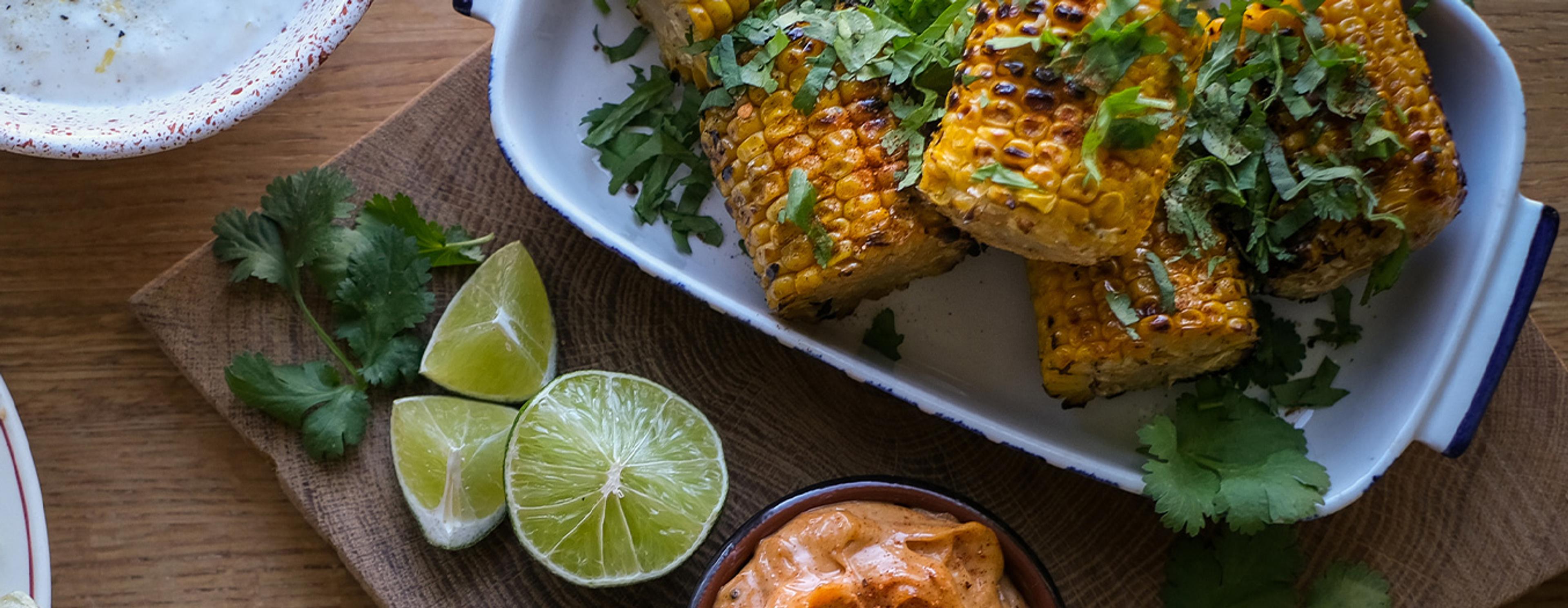 Grillad majs toppad med färska örter och limeklyftor på en träskärbräda, bredvid en fräsch sallad med krämig ost och rostade nötter. En krämig dippsås finns också med i bilden.