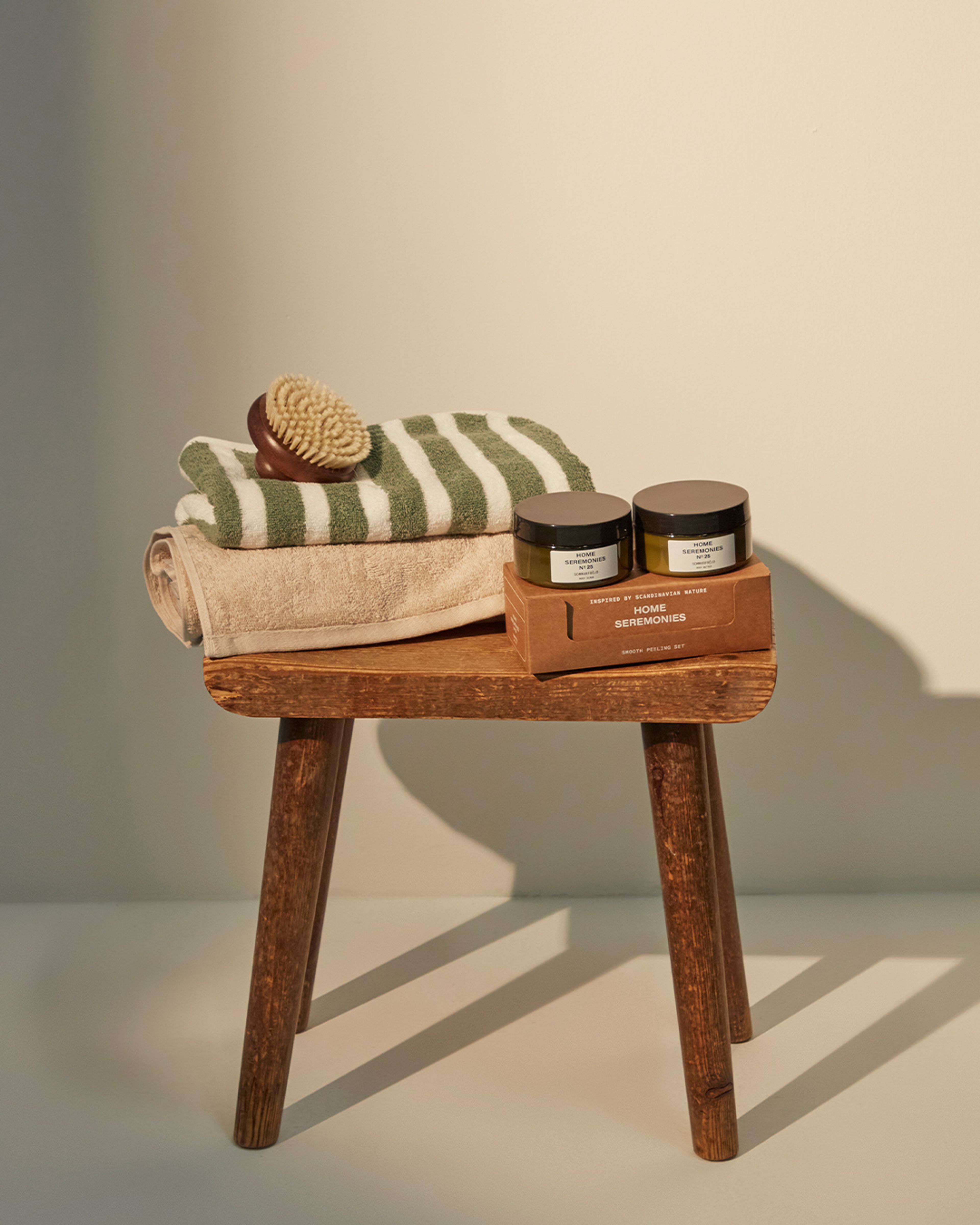 Ett set för hudpeeling med två burkar, en mjuk naturborste och två handdukar i beige och grönt, placerade på en enkel träpall.