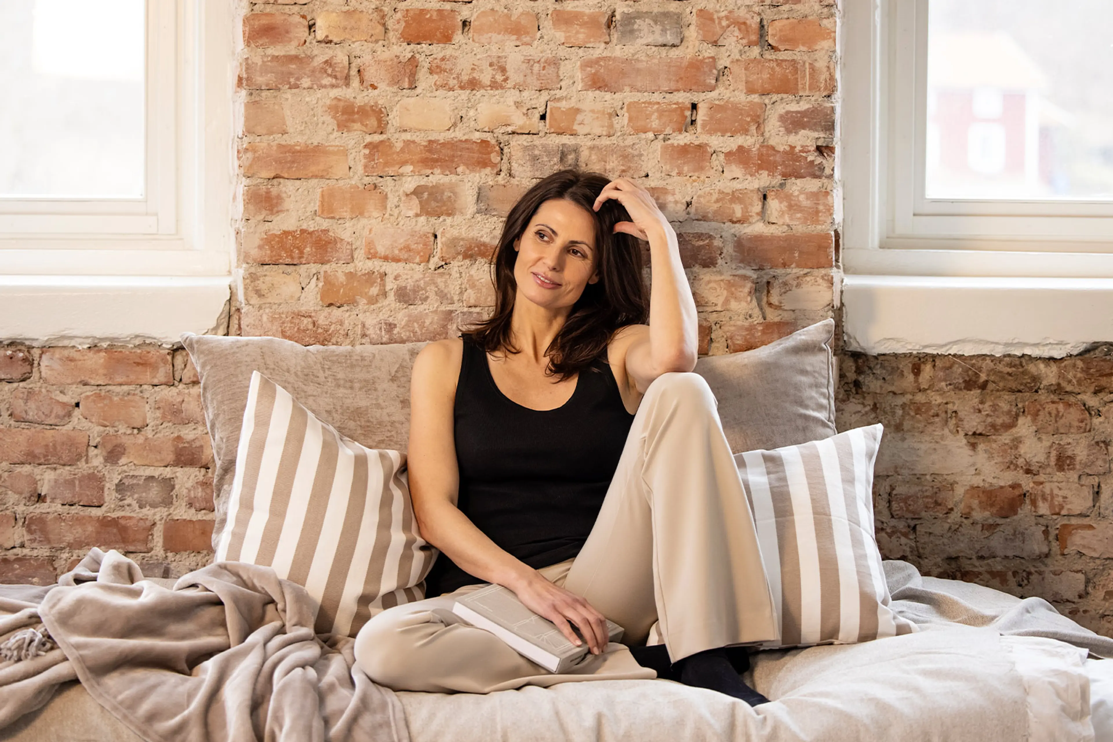 En svart ribbstickad linne-top i enkel och mjuk design, bärs av en kvinna som sitter avslappnat på en bädd med ljusa kuddar och plädar.