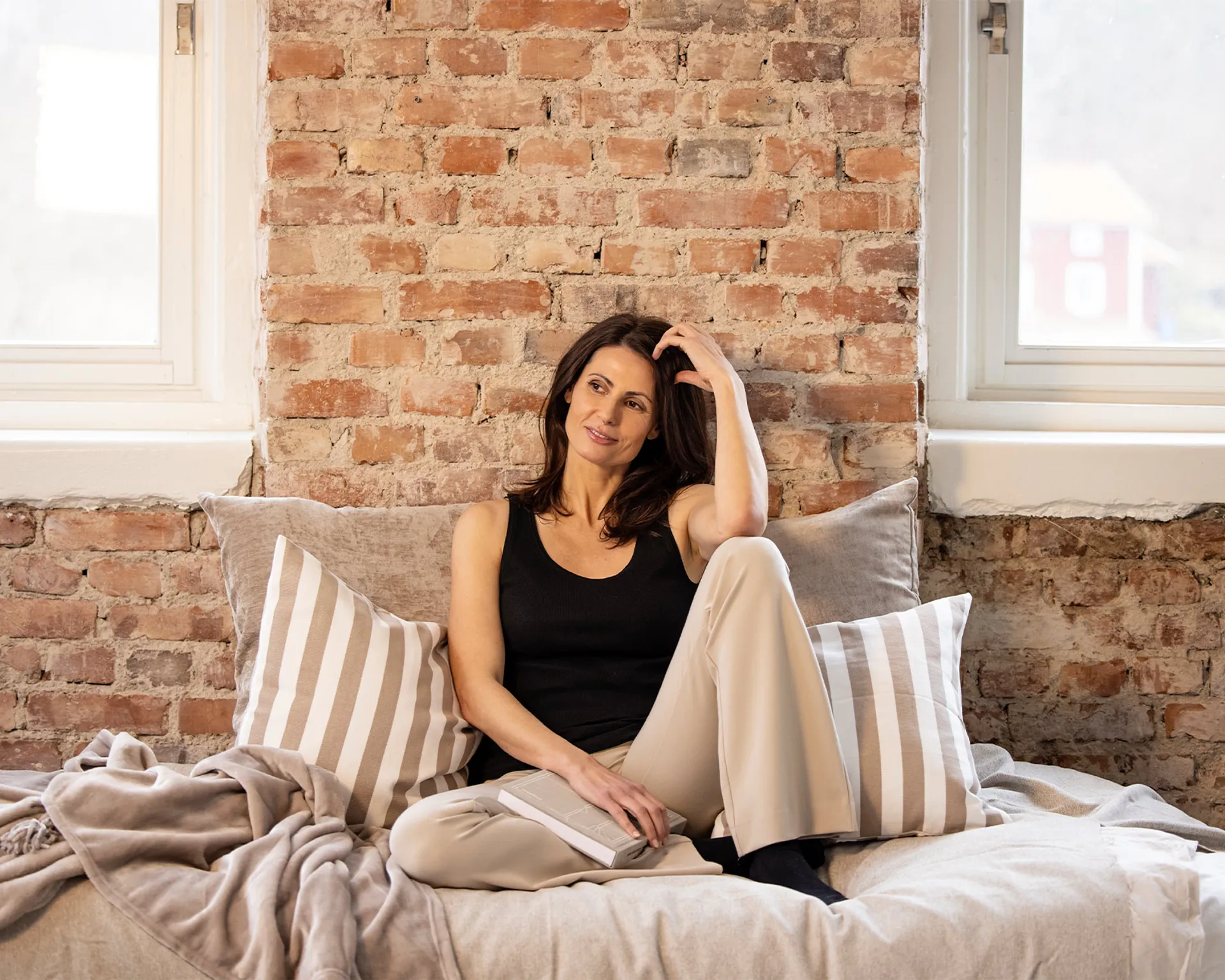 En svart ribbstickad linne-top i enkel och mjuk design, bärs av en kvinna som sitter avslappnat på en bädd med ljusa kuddar och plädar.
