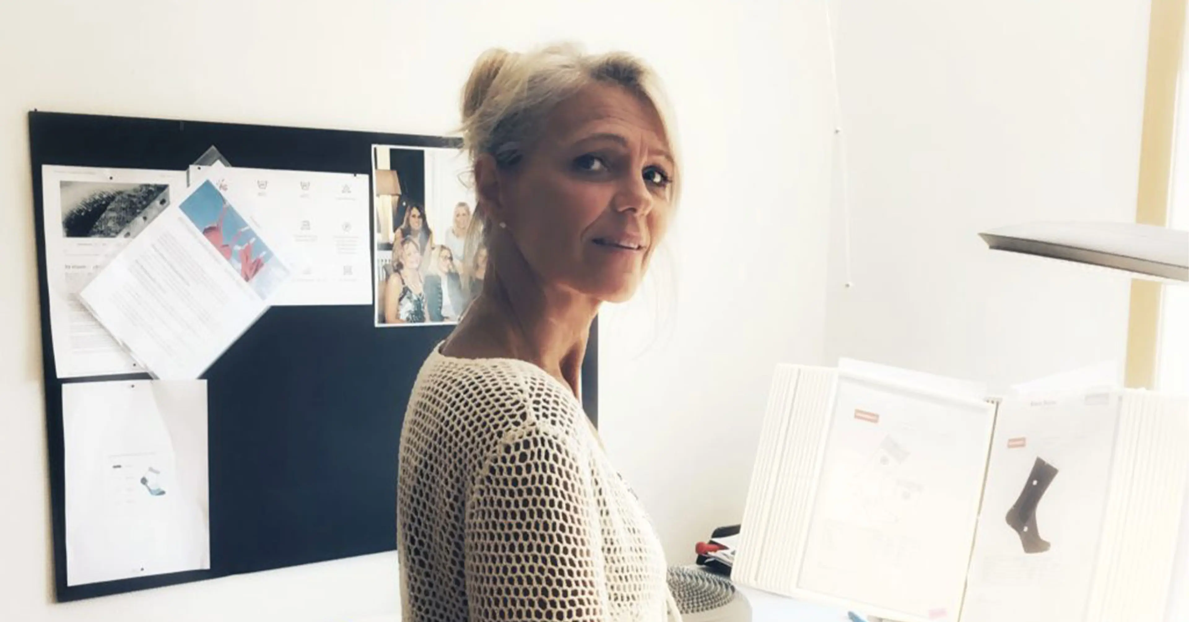 En kvinna i vit, hålmönstrad tröja arbetar vid ett upplyst skrivbord med dokument och ett par svarta strumpor. Bakom henne syns anslagstavla med papper och bilder.