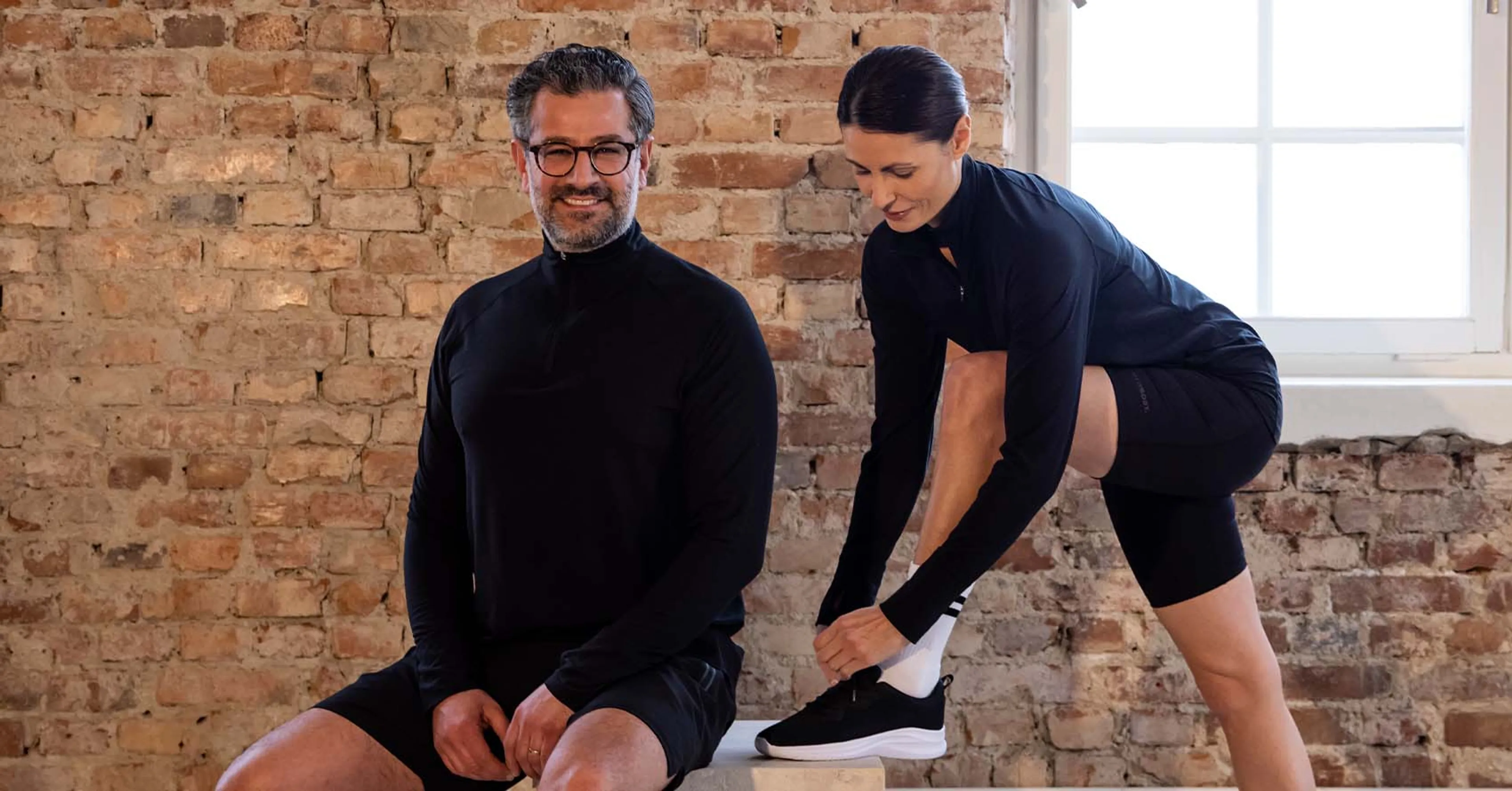 En man och en kvinna bär svarta långärmade tröjor med dragkedja, den ena står och ler medan den andra knyter sina skor. Bakgrunden är en vägg av rå tegelsten och ett stort fönster släpper in ljus.