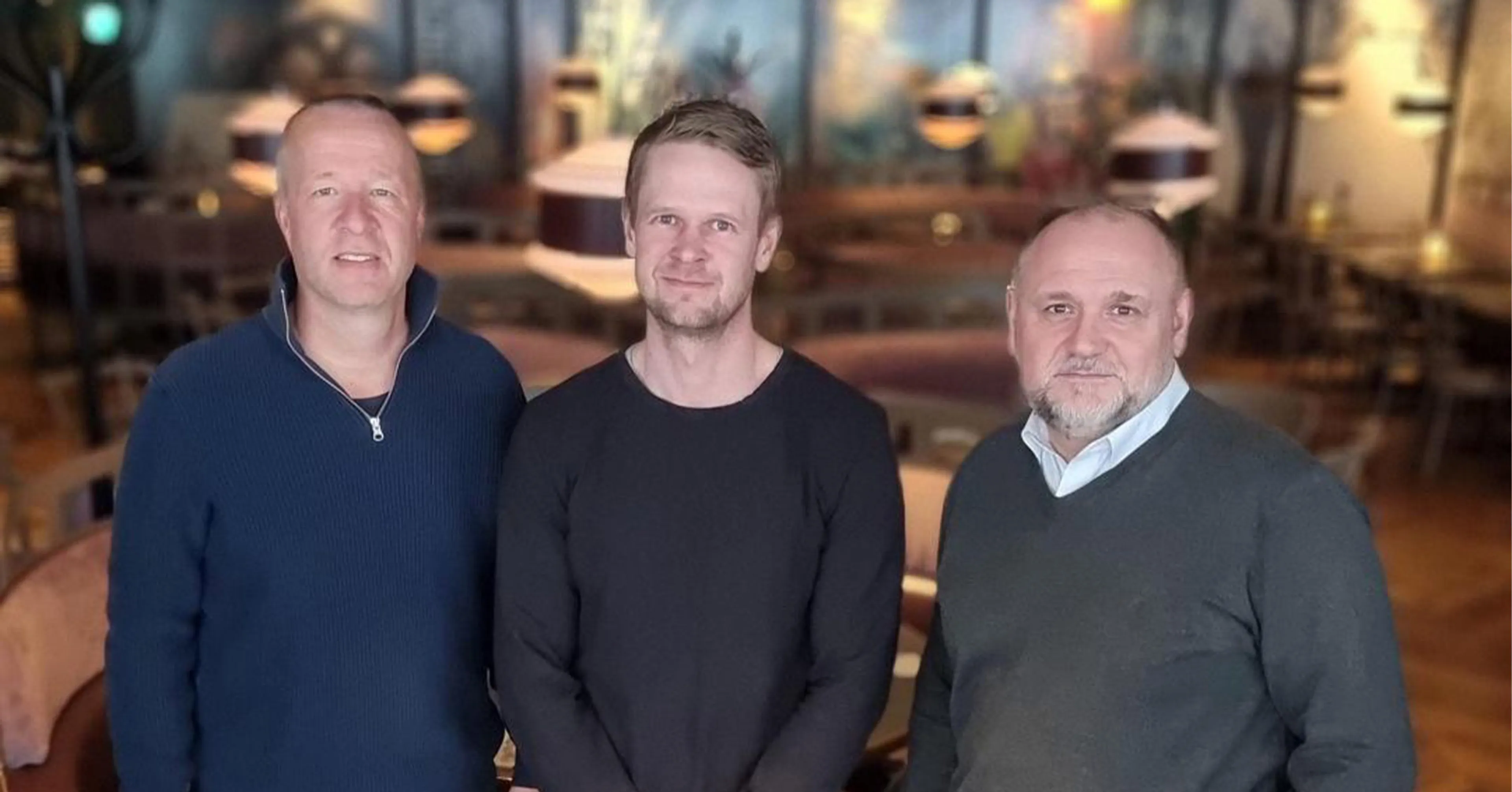 Tre män i tröjor står tillsammans i en mysig och varm inomhusmiljö med mjuk belysning och trämöbler runt omkring.