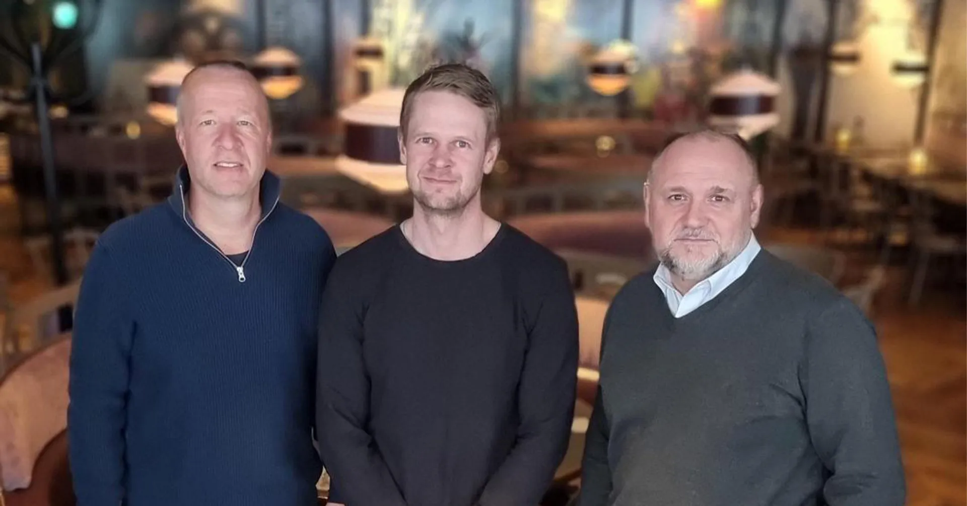Tre män i tröjor står tillsammans i en mysig och varm inomhusmiljö med mjuk belysning och trämöbler runt omkring.