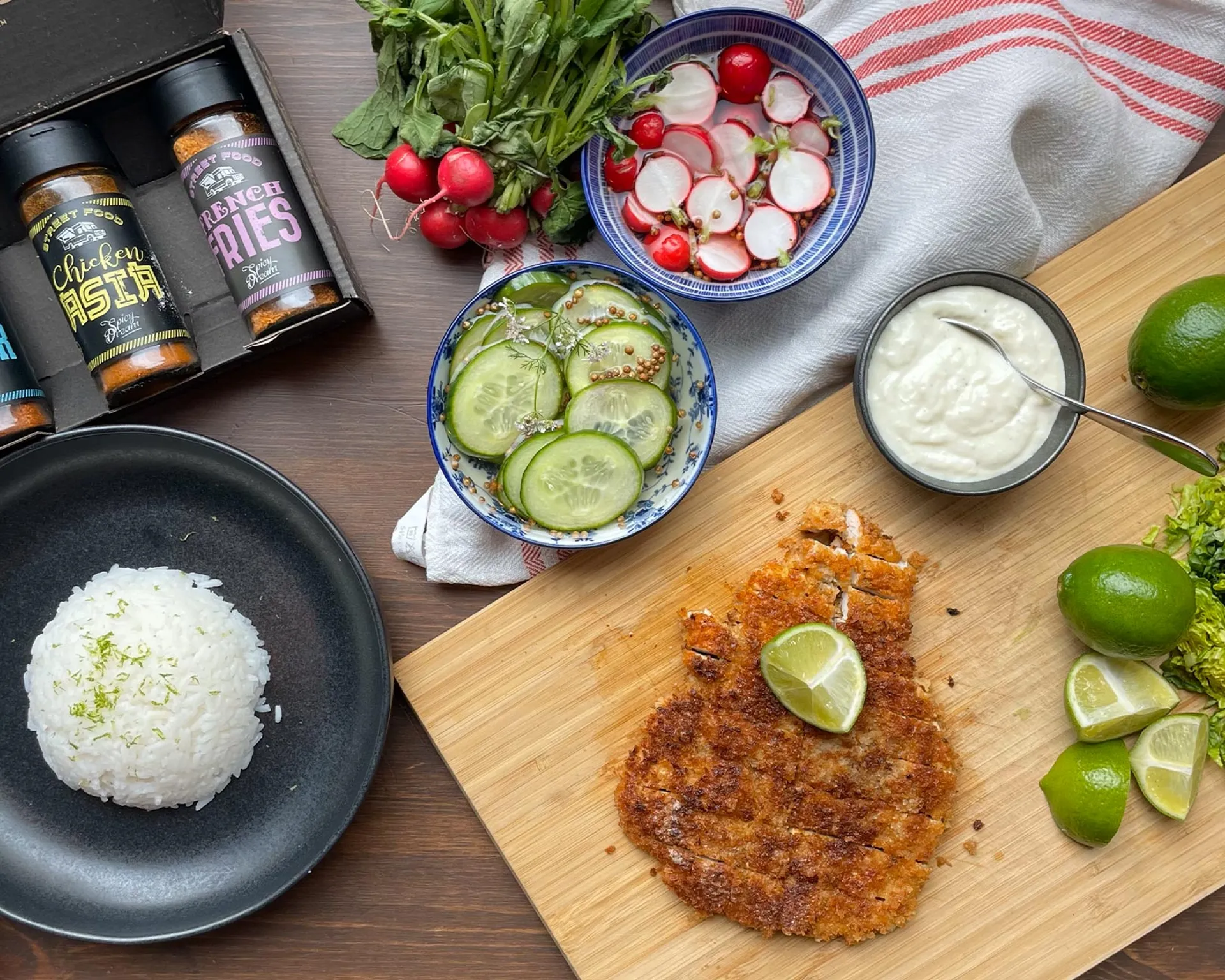 En krispig kycklingschnitzel med en limeklyfta på, serverad på ett skärbräde tillsammans med lime, en skål med sås och skivade grönsaker. På bordet finns också en portion ris och kryddburkar.