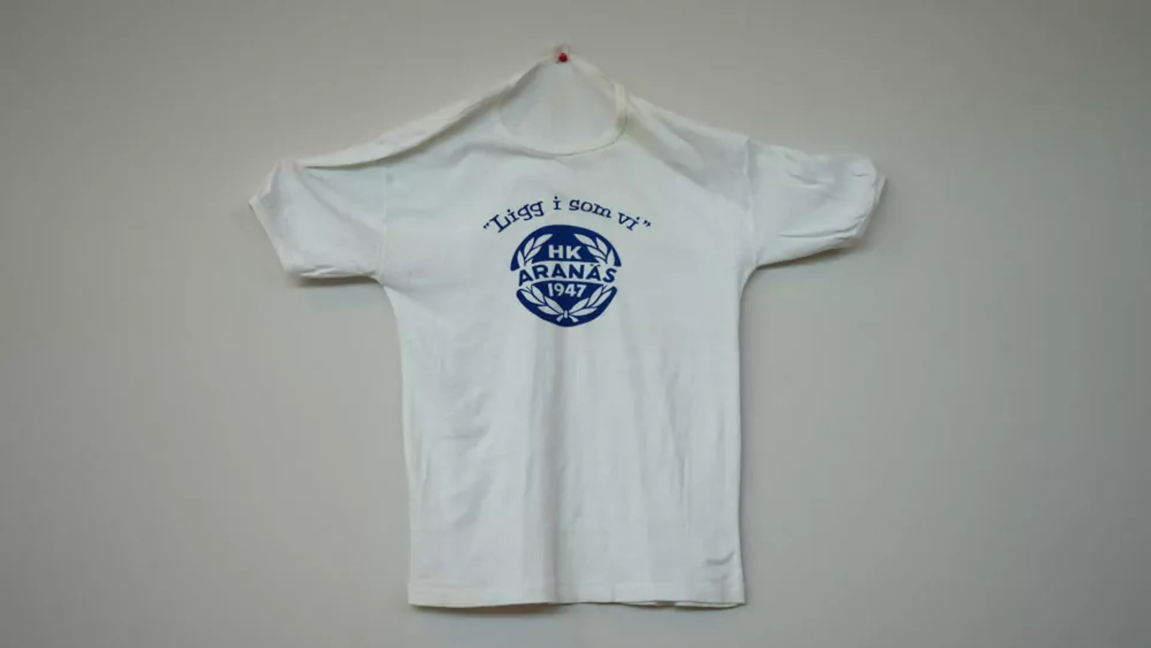 Vit kortärmad t-shirt med blå text och logotyp mitt på bröstet. Texten lyder "Liggg i som vi" och en emblemliknande symbol med "HK Aranäs 1947".