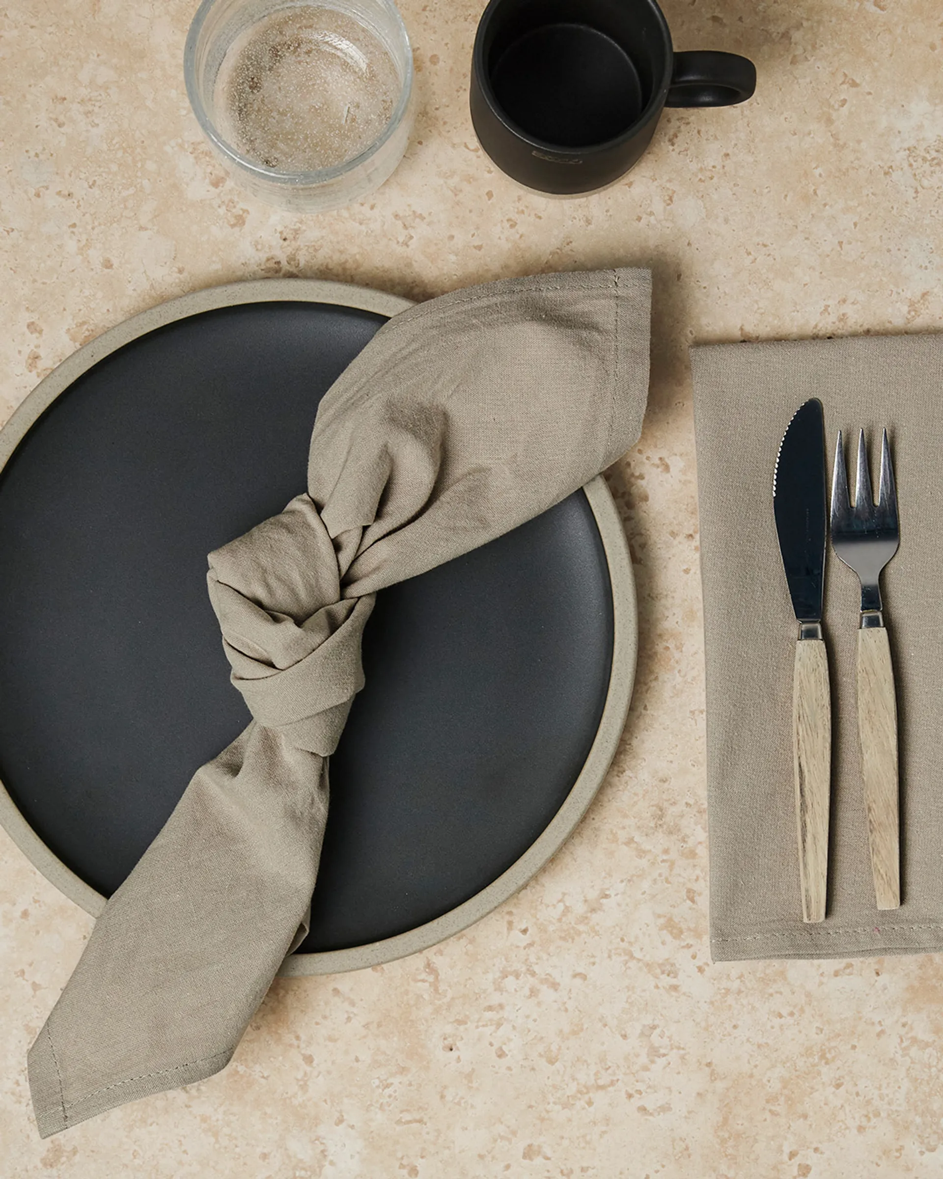 Snyggt dukat med en mörk tallrik och en knuten, beige tygservett. Bestick med trähandtag ligger på en matchande beige duk vid sidan om.