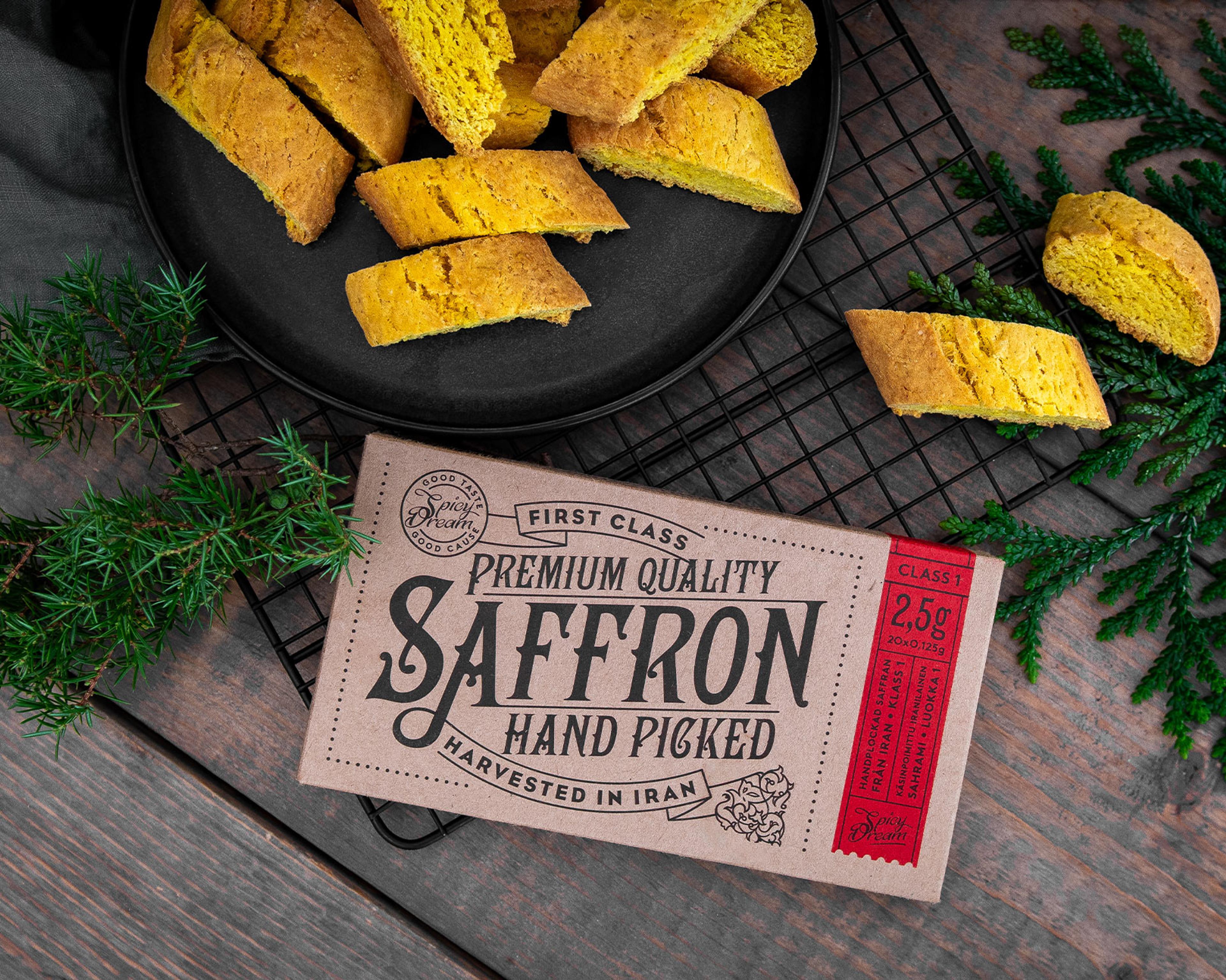 Ett paket med handplockad saffran från Iran ligger på ett bord bredvid flera bitar av gulbrunt bakverk på en svart plåt. Gröna kvistar pryder bilden för en naturlig känsla.