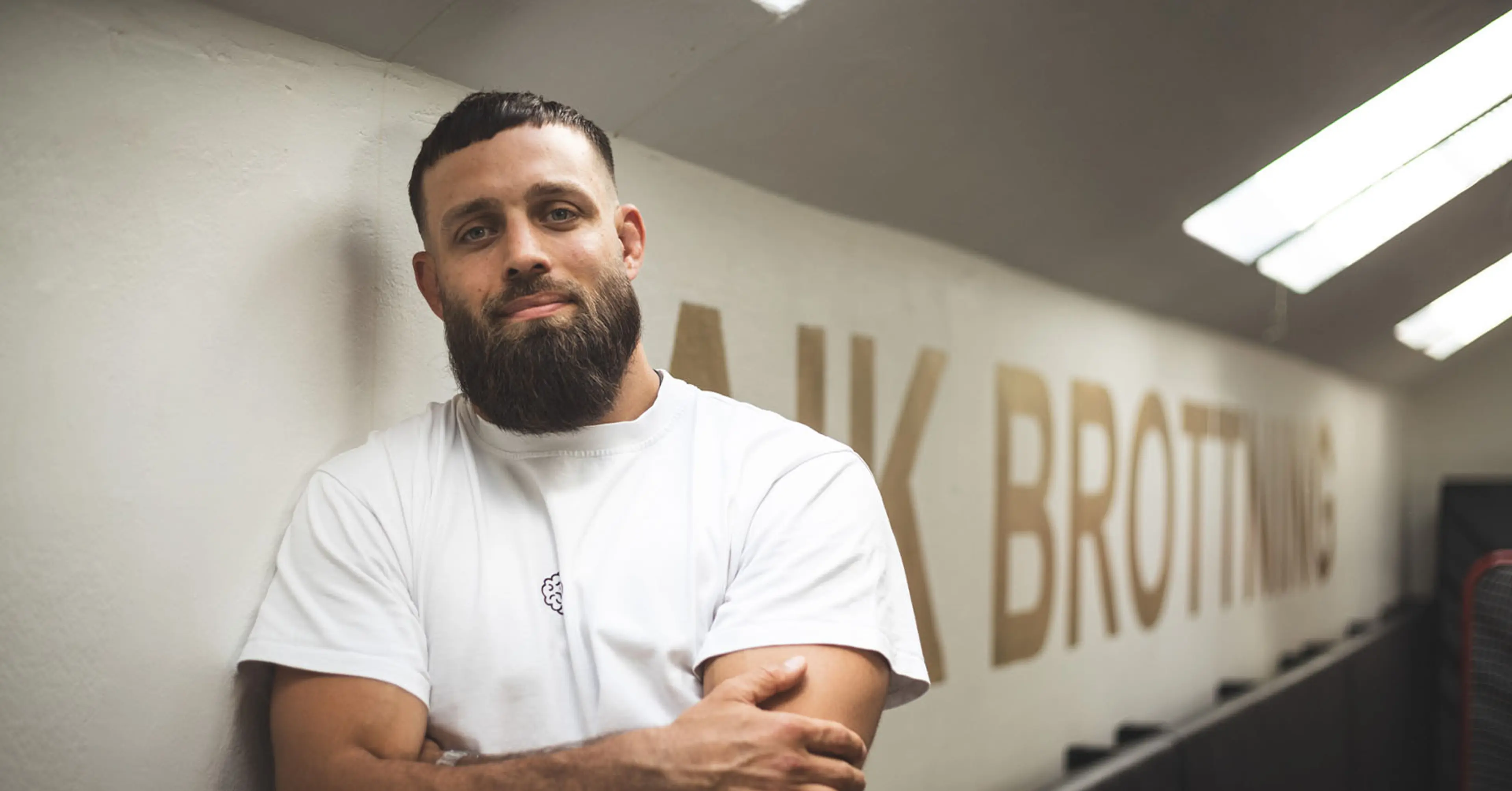 En man med skägg och vit t-shirt lutar avslappnat mot en vägg med texten “Brottning”. Ljuset från taklamporna skapar en mjuk atmosfär i träningslokalen.