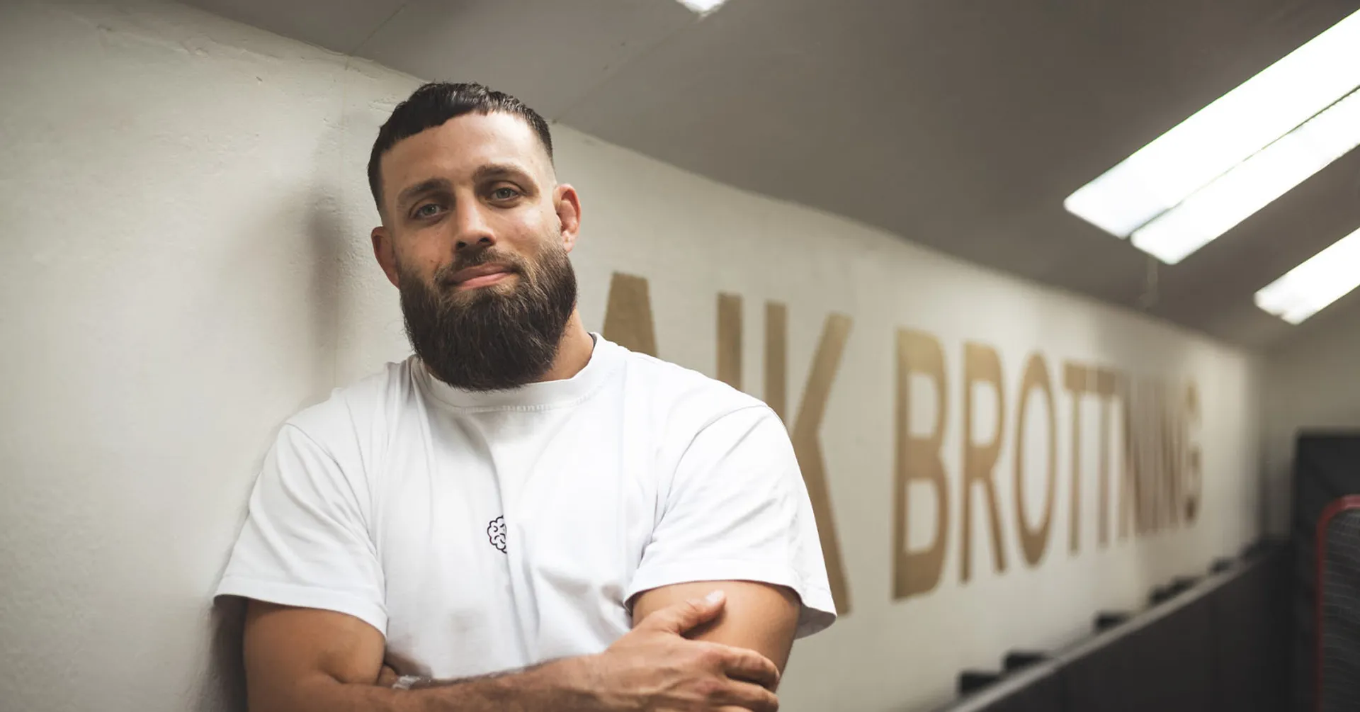 En man med skägg och vit t-shirt lutar avslappnat mot en vägg med texten “Brottning”. Ljuset från taklamporna skapar en mjuk atmosfär i träningslokalen.