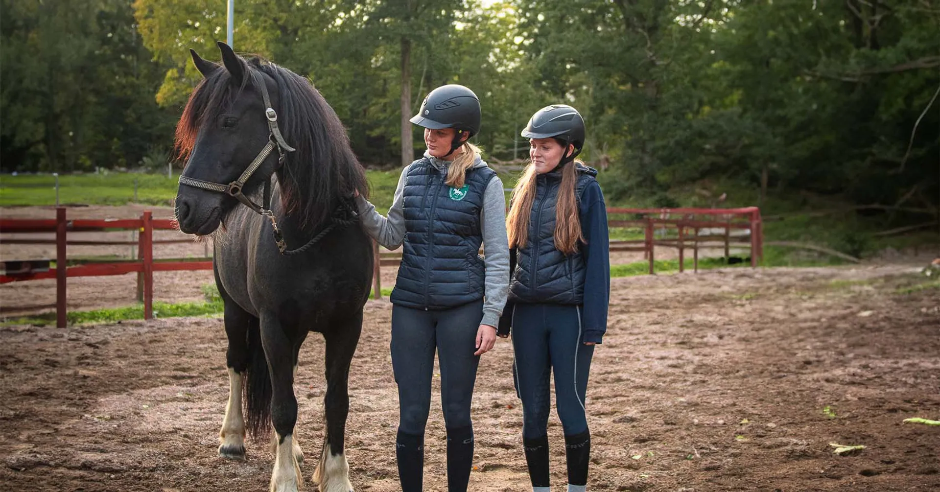 Två tjejer med ridhjälm som står bredvid en mörk häst men lång mörk man
