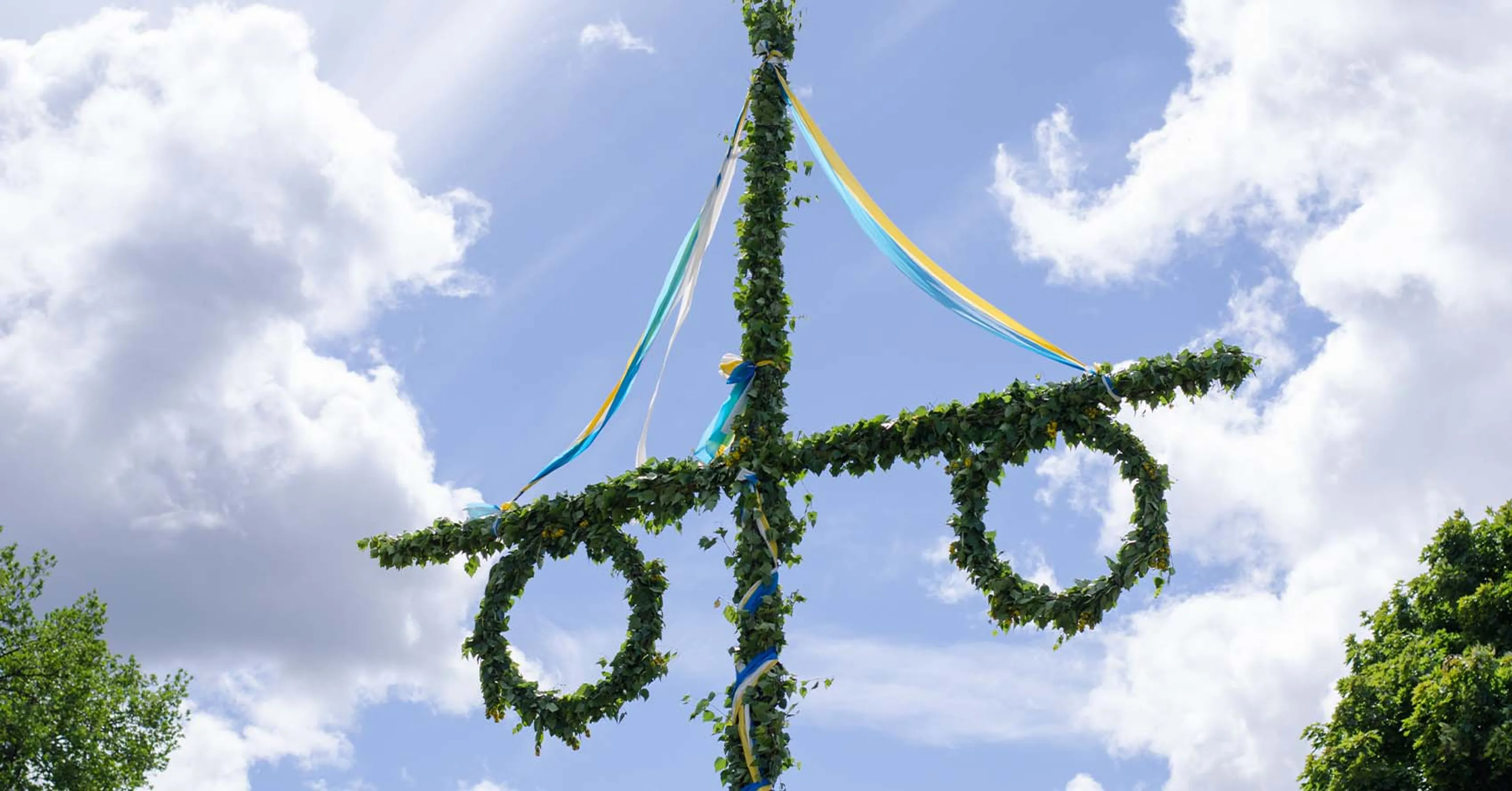 En traditionell midsommarstång dekorerad med gröna kvistar och slingor, omgiven av frodig grönska under en blå himmel. En svensk flagga vajar bredvid i lätt sommarbris.