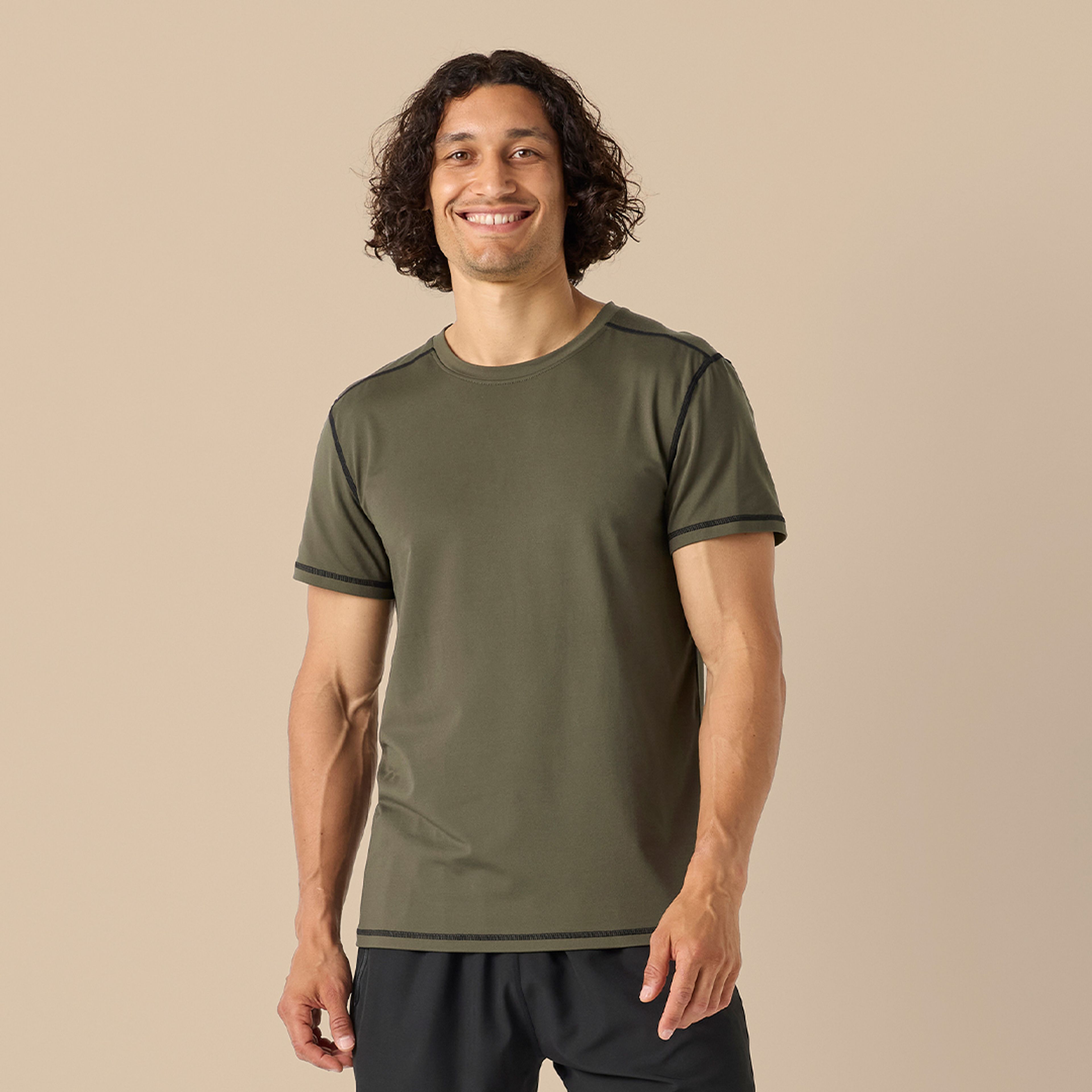 En kortärmad t-shirt i olivgrönt med mörka kontrastsömmar i en sportig och funktionell design. Bärs av en leende person mot en neutral bakgrund.