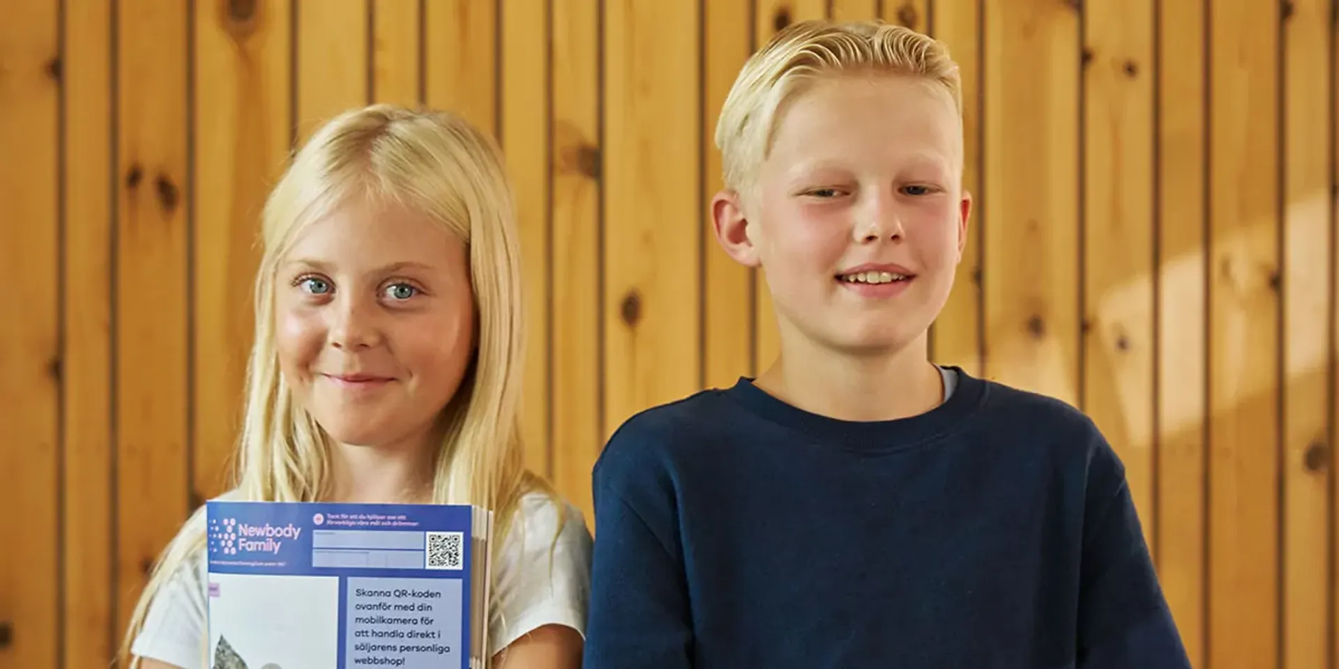 Två barn står framför en trävägg och håller i en försäljningskatalog med QR-kod för smidig beställning. De ser glada och stolta ut över sitt uppdrag att tjäna pengar tillsammans.