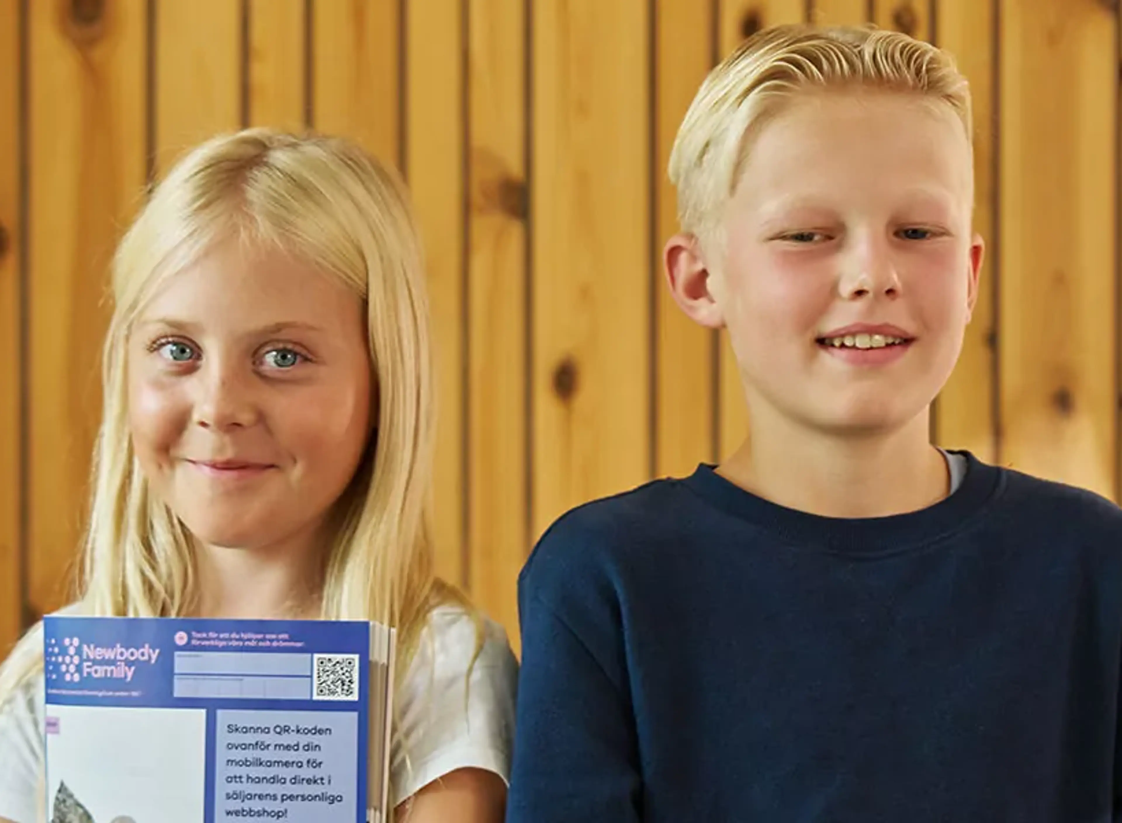 Två barn står framför en trävägg och håller i en försäljningskatalog med QR-kod för smidig beställning. De ser glada och stolta ut över sitt uppdrag att tjäna pengar tillsammans.