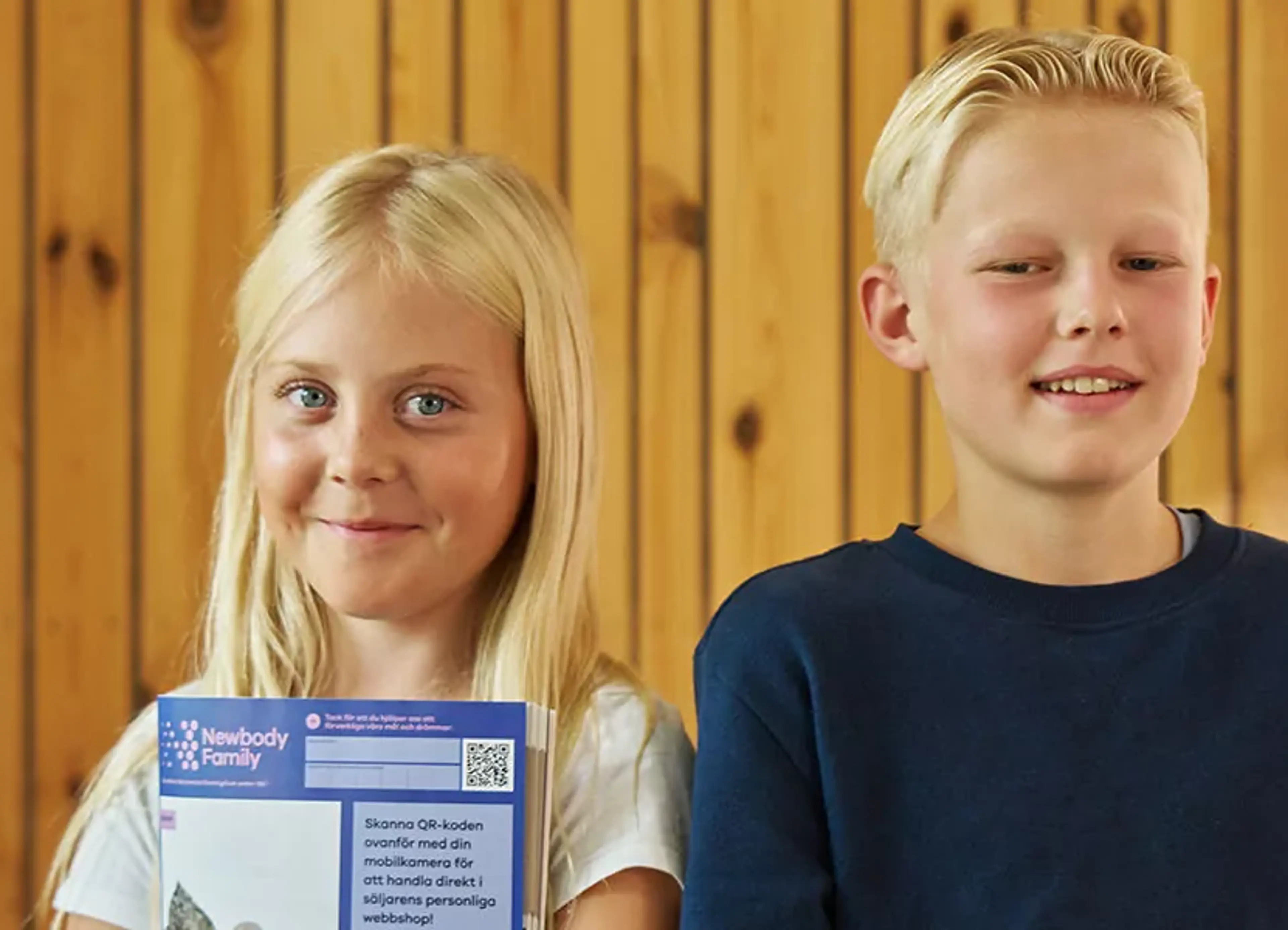 Två barn står framför en trävägg och håller i en försäljningskatalog med QR-kod för smidig beställning. De ser glada och stolta ut över sitt uppdrag att tjäna pengar tillsammans.