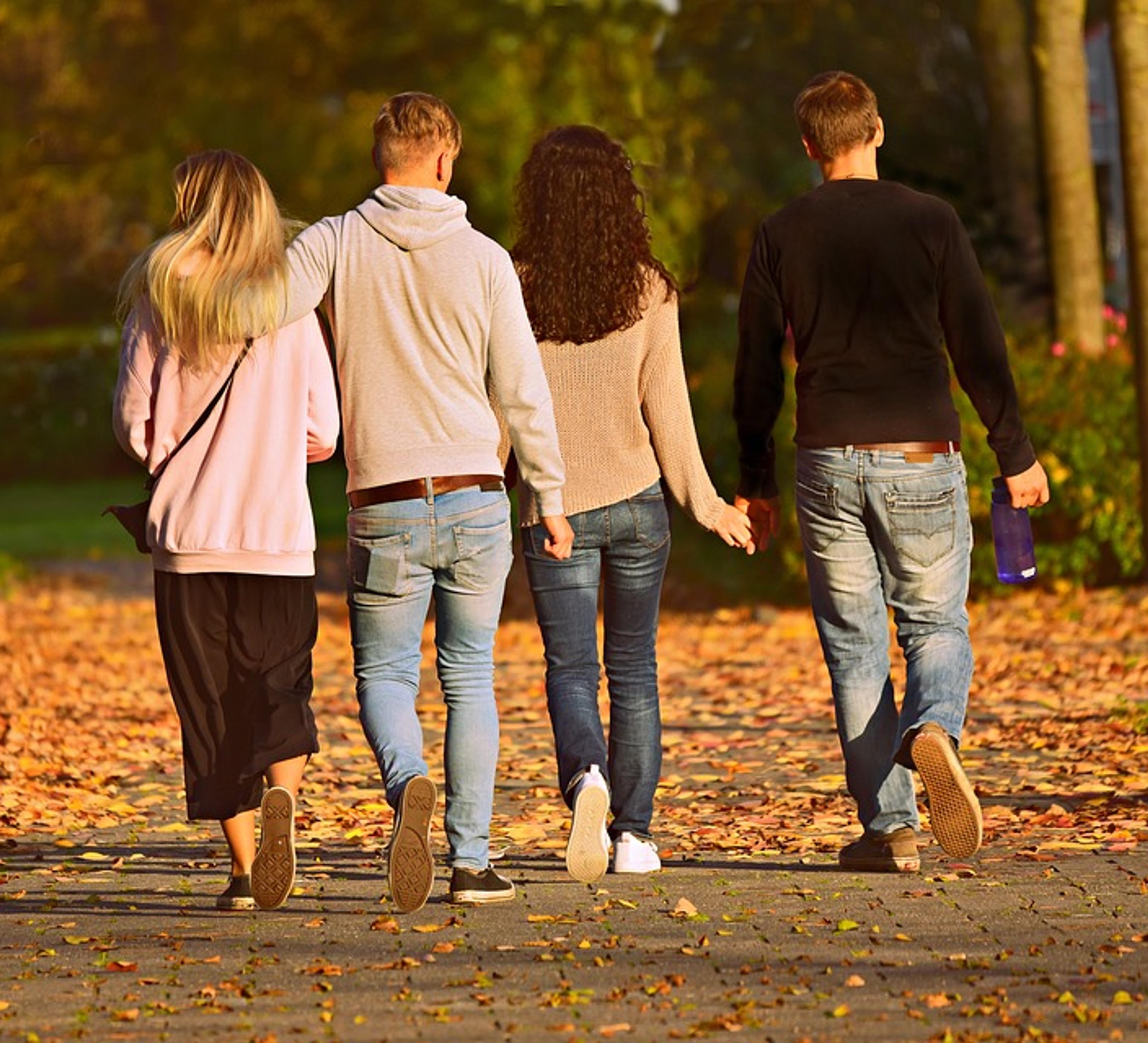 Fyra personer går tillsammans på en lövbeklädd stenlagd stig bland träd på hösten. Två håller varandra i handen medan de promenerar bort från kameran.